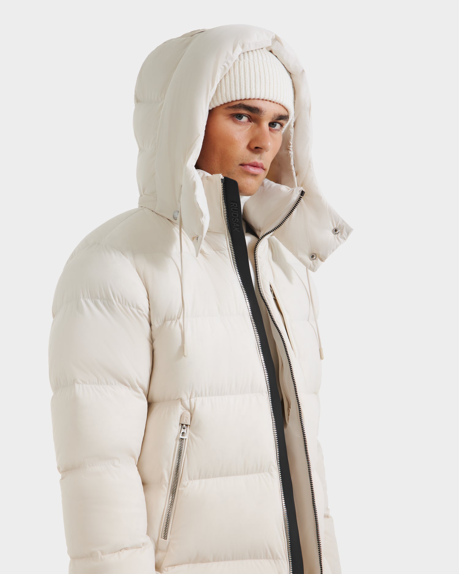 A person wearing the Rudsak VINCE M - 6124561 Vanilla mens quilted down puffer jacket with a hood and a white knit beanie, looking slightly to the side against a plain white background. The jacket features RDS-certified down fill.