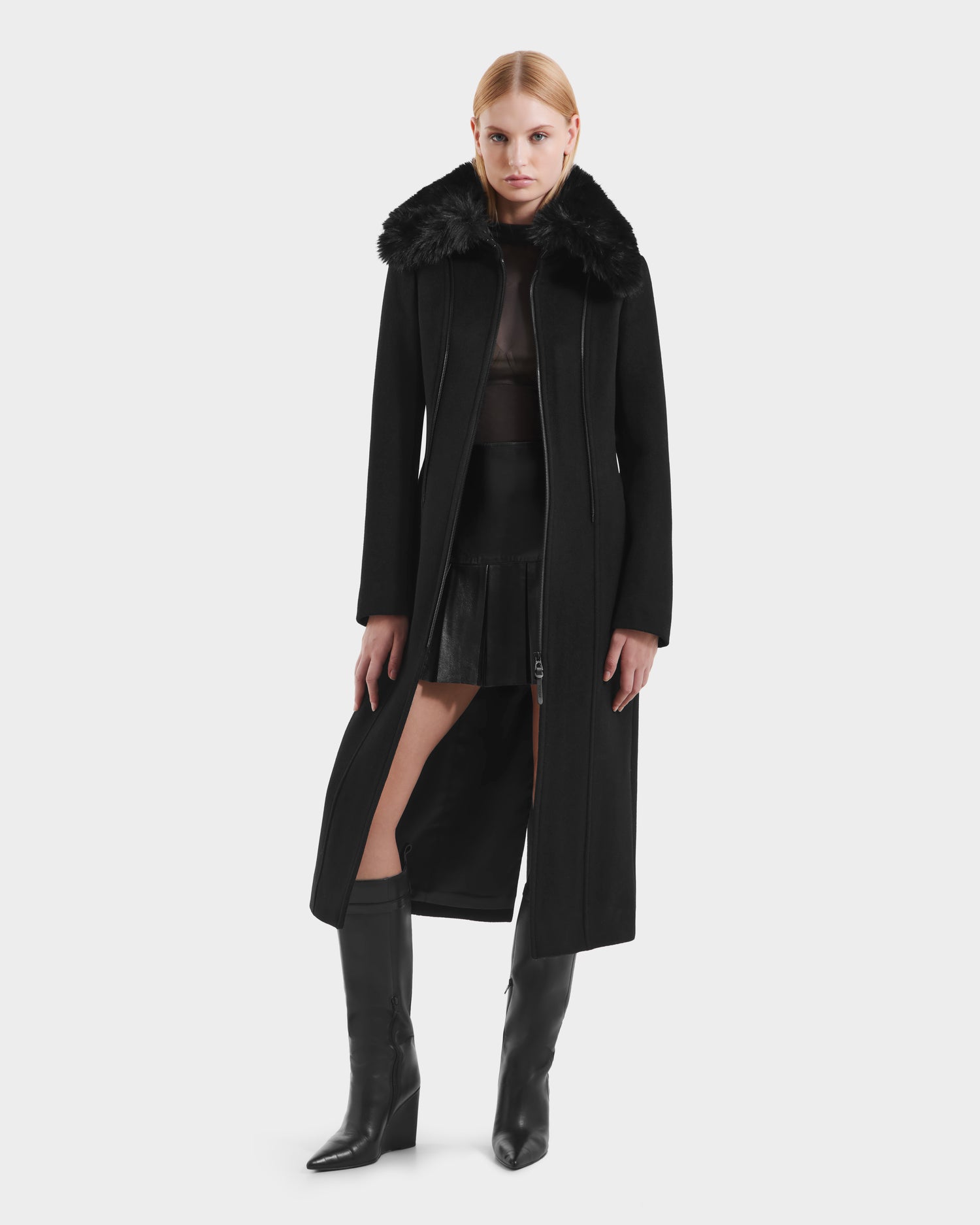 A woman stands against a white background wearing the VIRGINIE - 8125698 Black women’s long peacoat with a removable faux fur collar by Rudsak, paired with a sheer black top, short black skirt, and knee-high black boots.