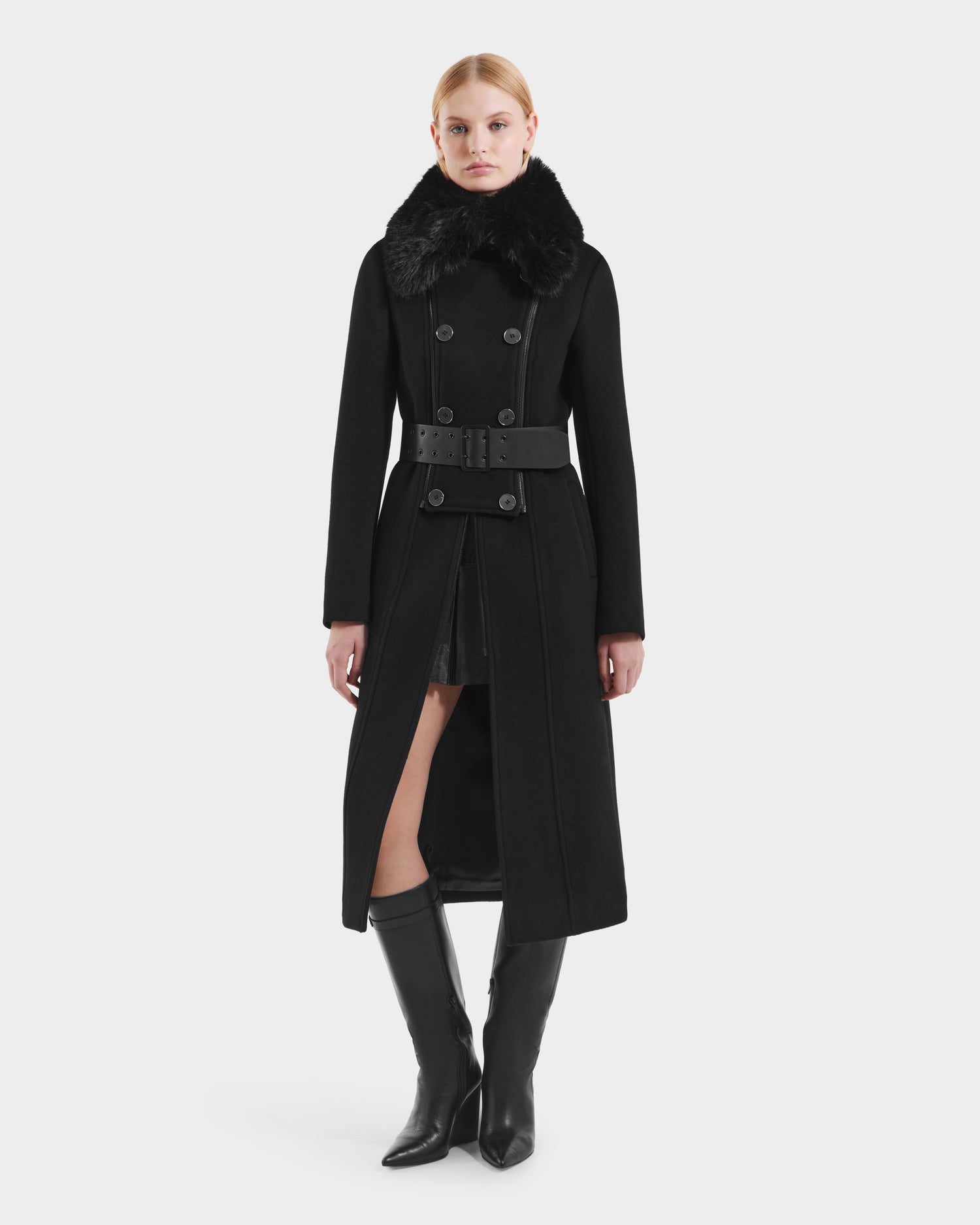 A woman faces forward in the Rudsak VIRGINIE - 8125698 Black peacoat: a black wool blend with removable faux fur collar and matching belt, partially open at the bottom to reveal a black outfit, paired with tall black boots.