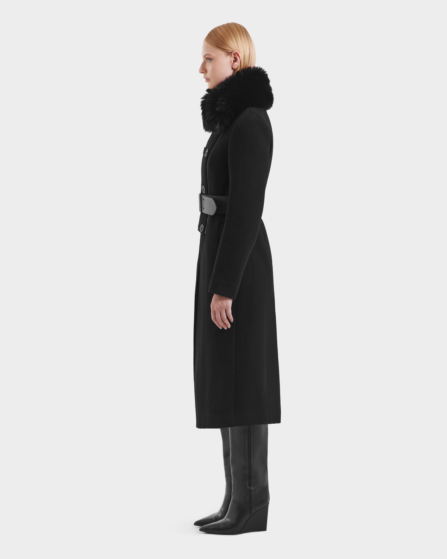 A woman with straight blonde hair stands in profile, wearing the Rudsak VIRGINIE - 8125698 Black womens long peacoat with a removable faux fur collar and belted waist, paired with tall black boots against a plain white background.