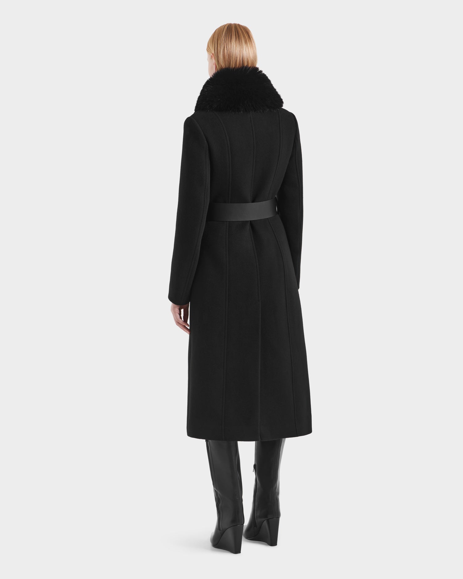 A blonde woman stands facing away, wearing the Rudsak VIRGINIE - 8125698 Black long peacoat with a removable faux fur collar and wide belt, paired with black knee-high boots.