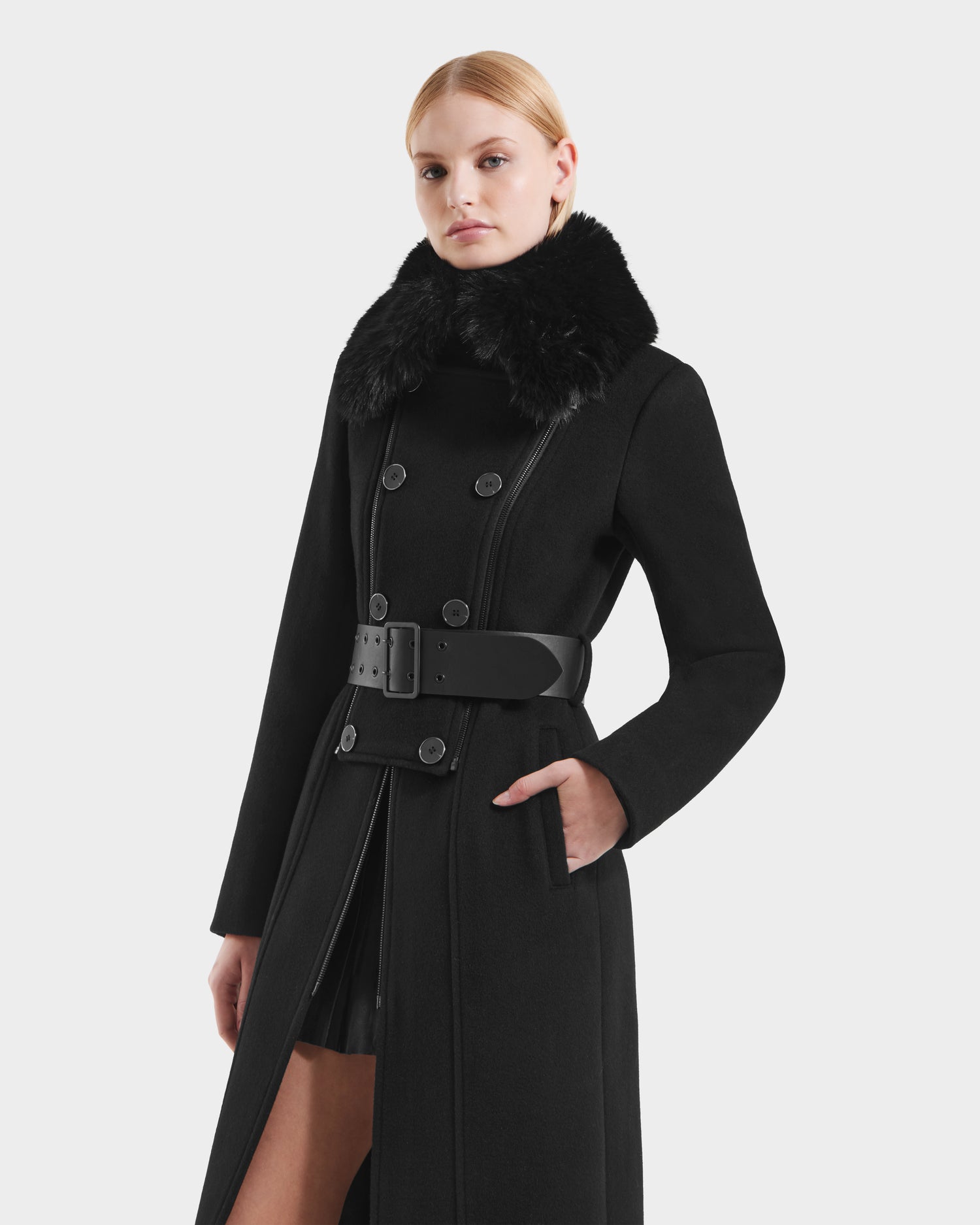 A woman stands with one hand in her pocket wearing Rudsaks VIRGINIE - 8125698 Black women’s long peacoat, featuring a wide belt and removable faux fur collar, against a plain white background.