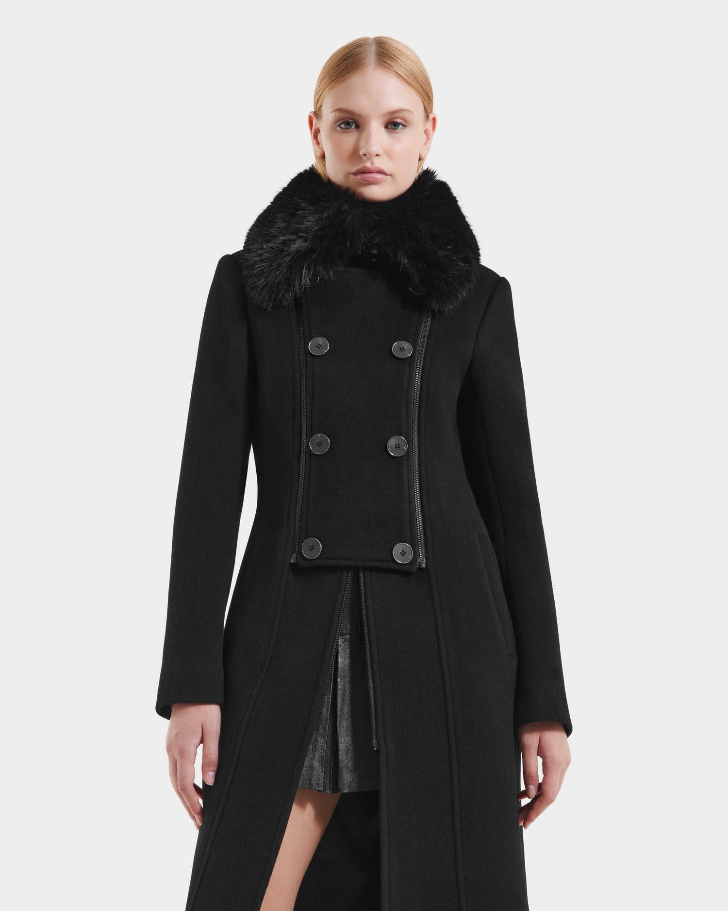 A woman wearing the Rudsak VIRGINIE - 8125698 Black womens long peacoat with large black buttons and a wide removable faux fur collar stands against a plain white background.