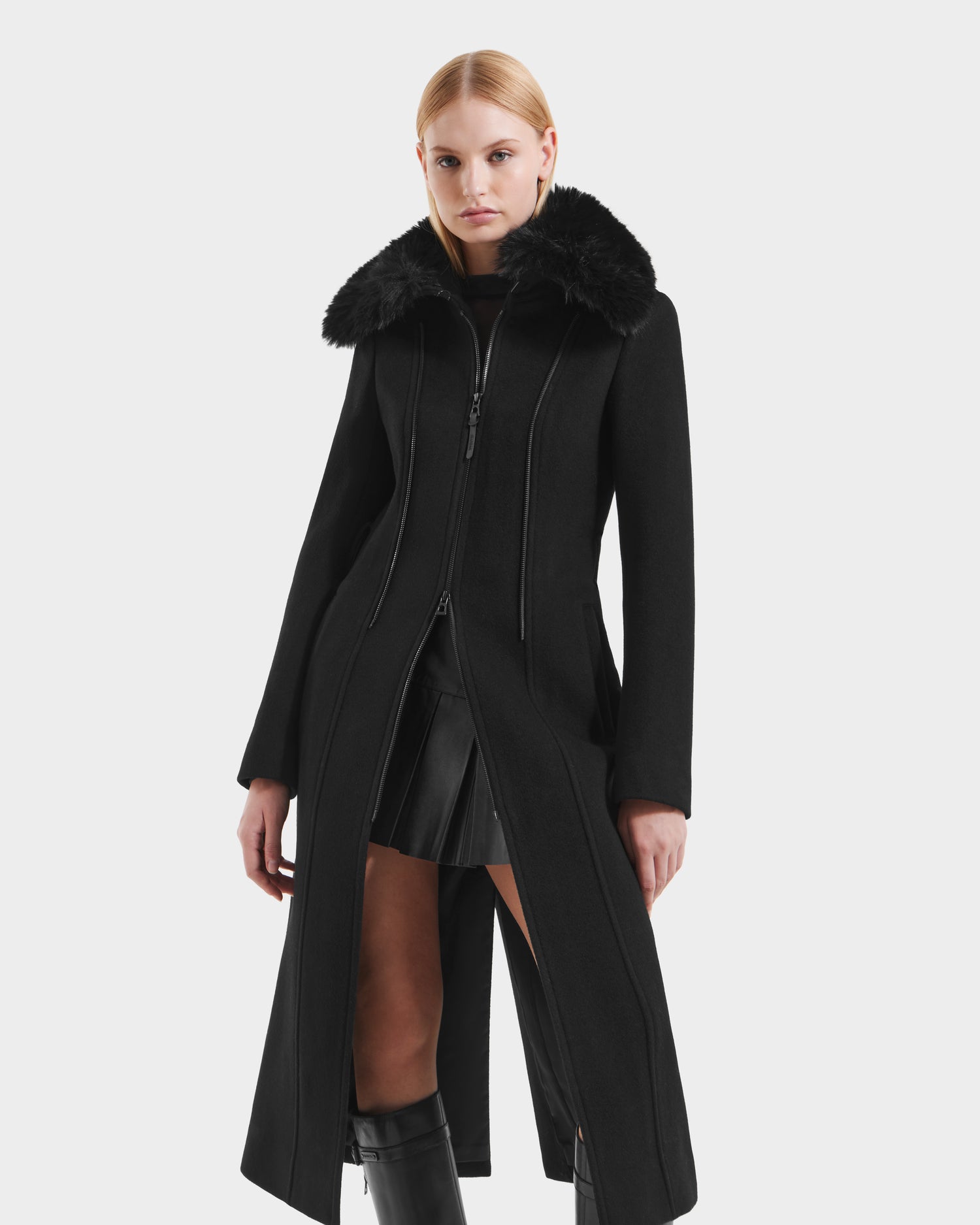 A person wearing the Rudsak VIRGINIE - 8125698 Black womens long peacoat with a removable faux fur collar and zippered front stands against a plain light background, paired with a black pleated skirt.
