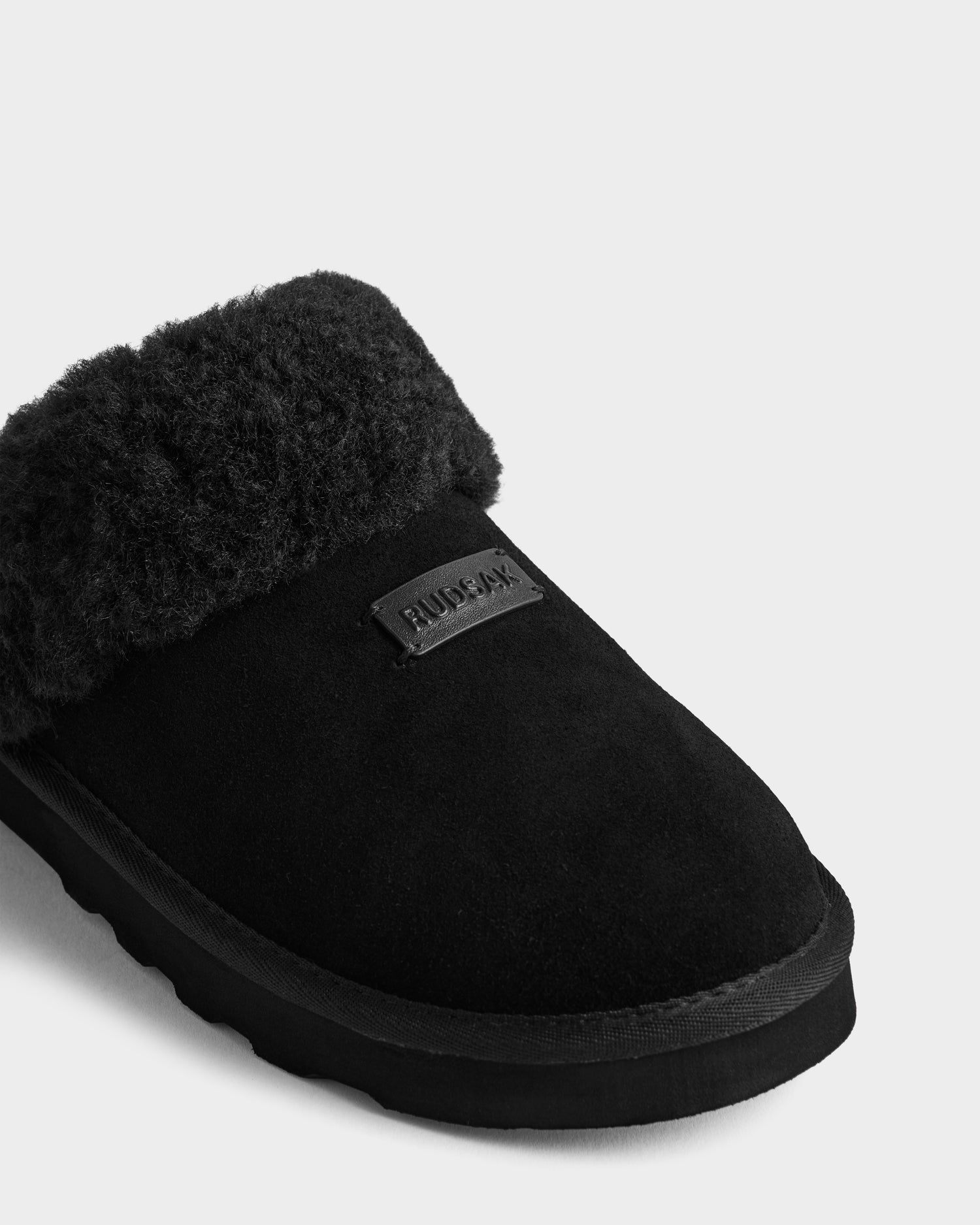 A close-up of the Rudsak WILBER - 6225509 Black men’s suede slipper with a soft faux shearling cuff and Rudsak label on the upper, shown on a light background—ideal for elevated loungewear style.