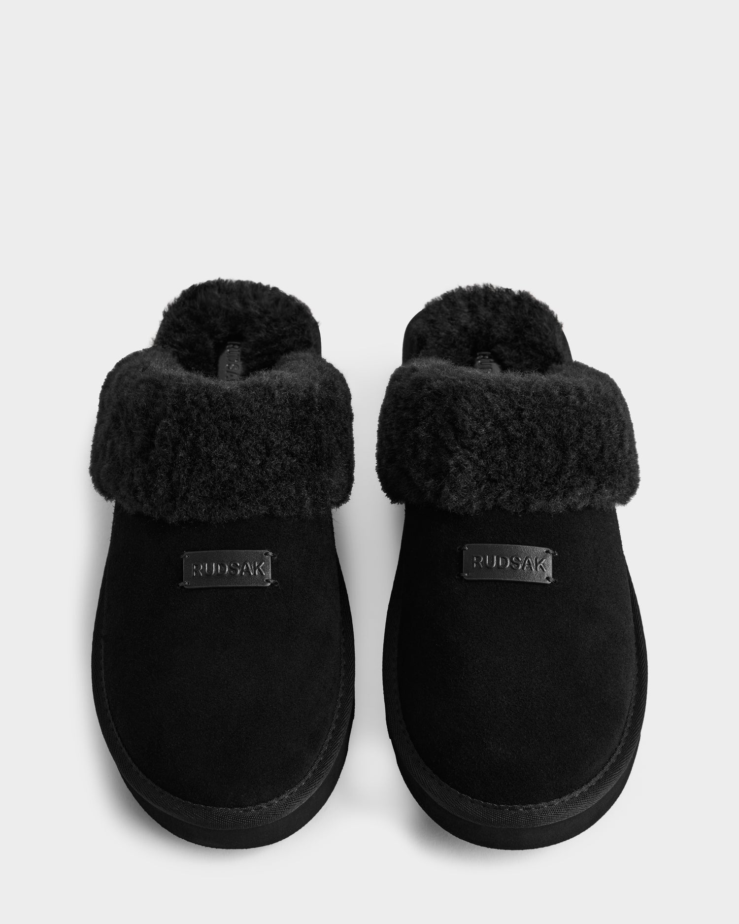 The Rudsak WILBER - 6225509 Black men's slippers feature plush faux shearling cuffs, a suede-like finish, and RUDSAK branding—ideal for elevating your loungewear style.