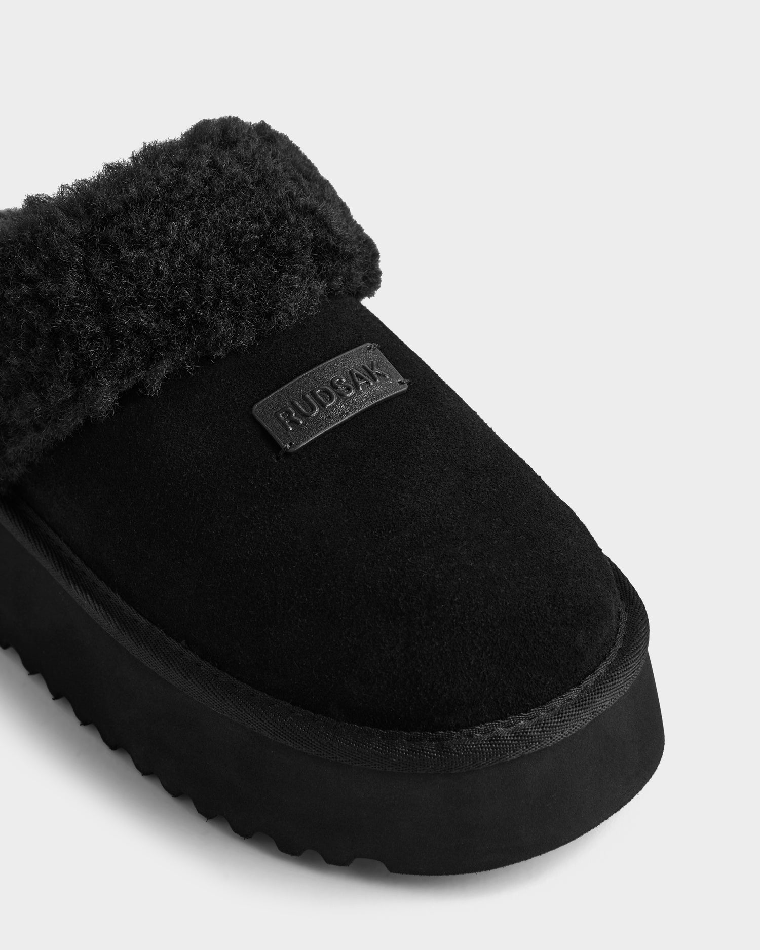 A close-up of the WILMA - 8225582 Black by Rudsak shows a black suede women’s slipper with a fuzzy lining, textured sole, and a stitched Rudsak patch—making it perfect for cozy winter fashion.