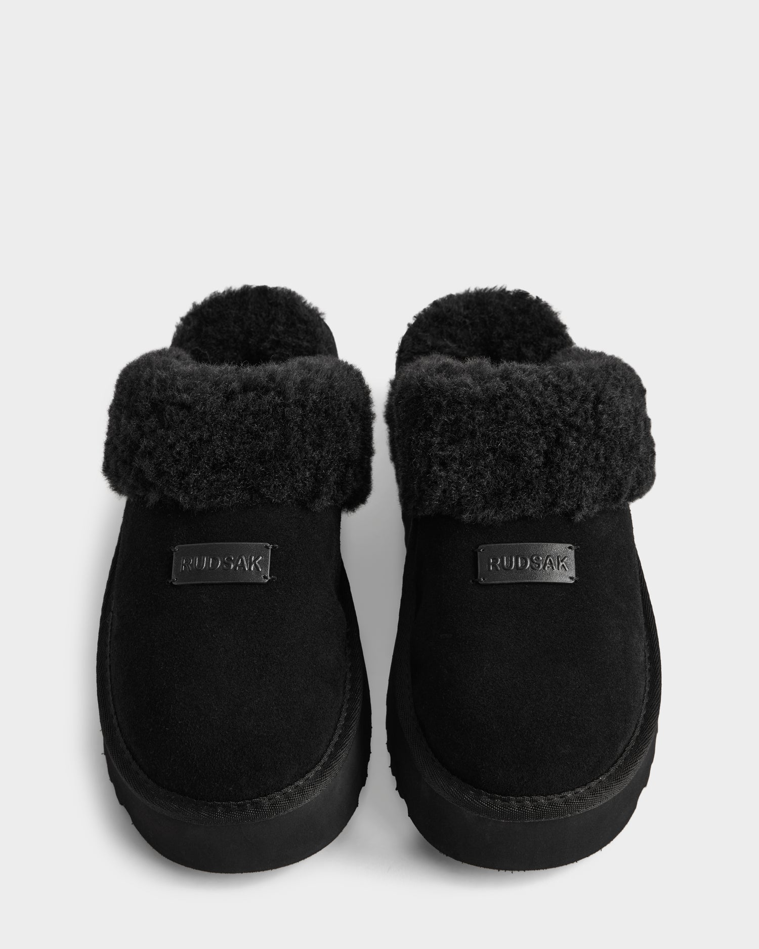 A pair of WILMA - 8225582 Black suede slipper slip-ons by Rudsak, with thick faux fur lining and fold-over cuffs, are shown side by side on a white background. The women’s slippers feature a front label reading RUDSAK—perfect for winter.