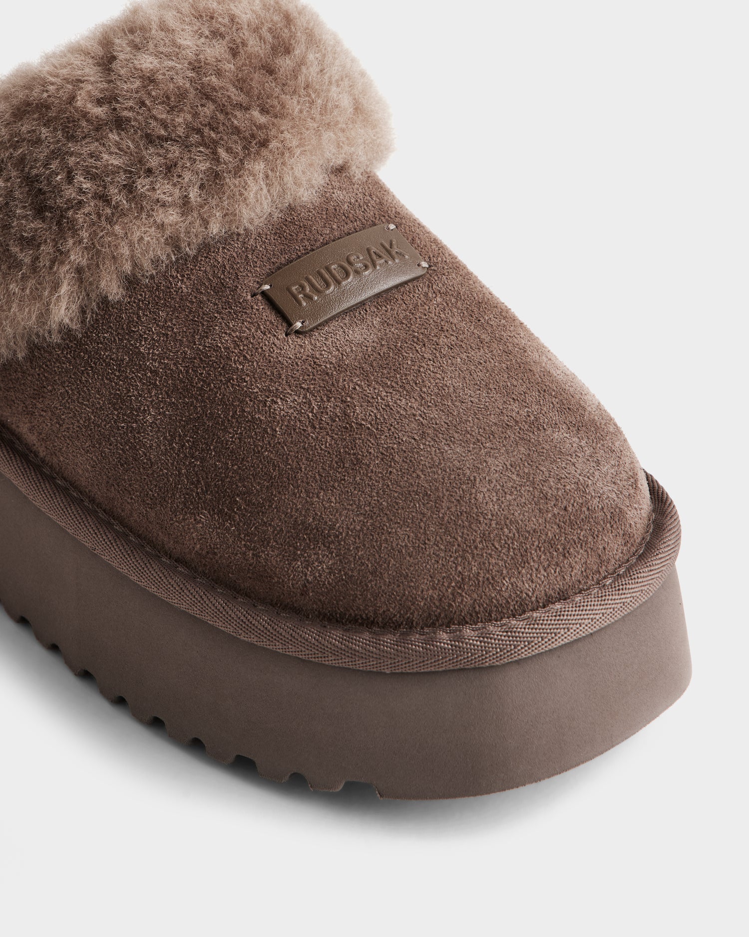 Close-up of the Rudsak WILMA - 8225582 Mocha suede women’s slipper, featuring a fuzzy lining, thick platform sole, and a rectangular Rudsak label stitched on top. Set against a white background.