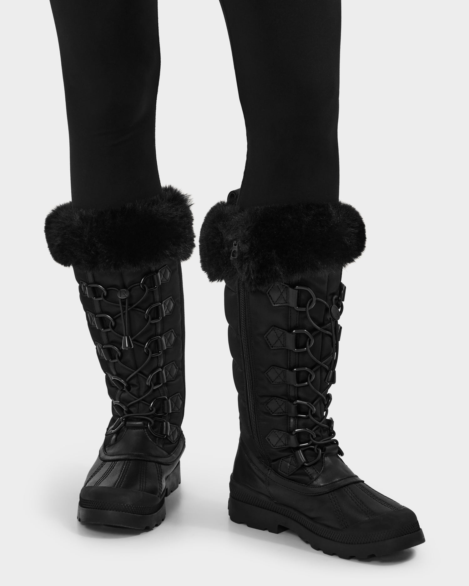 A person wears the Rudsak WINDY - L224507 Black women's winter boots with faux fur trim and decorative stitching, paired with black leggings. These boots feature rugged soles for cold-weather comfort and style.
