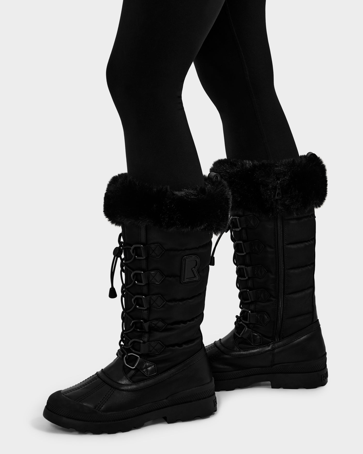 A person in black leggings wears the Rudsak WINDY - L224507 Black women's winter boots, featuring quilted sides, lace-up fronts, and cozy faux fur trim—perfect for cold-weather comfort—standing on a light background.