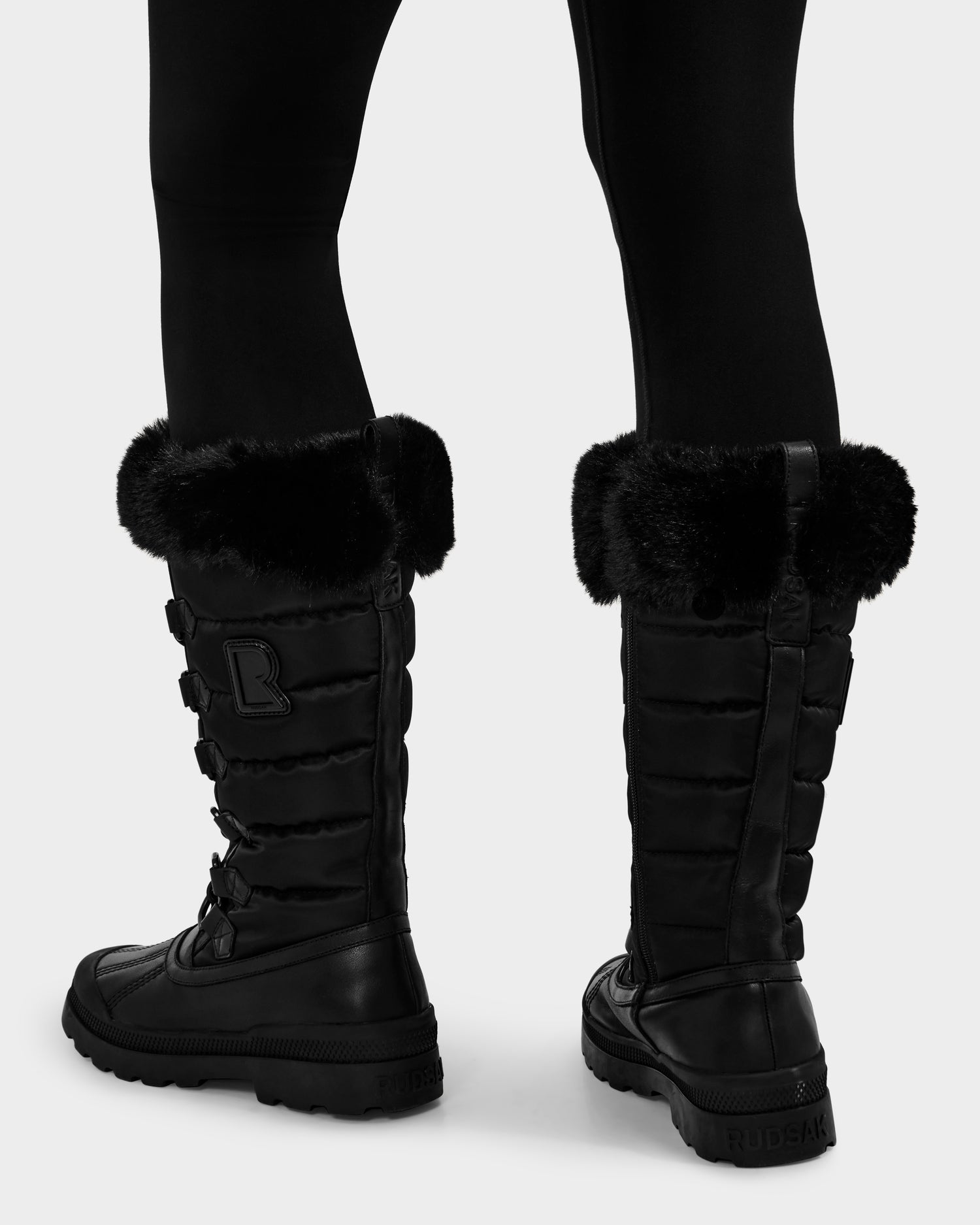 A person shown from the back is wearing Rudsak's WINDY - L224507 Black knee-high women's winter boots with faux fur trim and lace-up details, paired with black leggings.