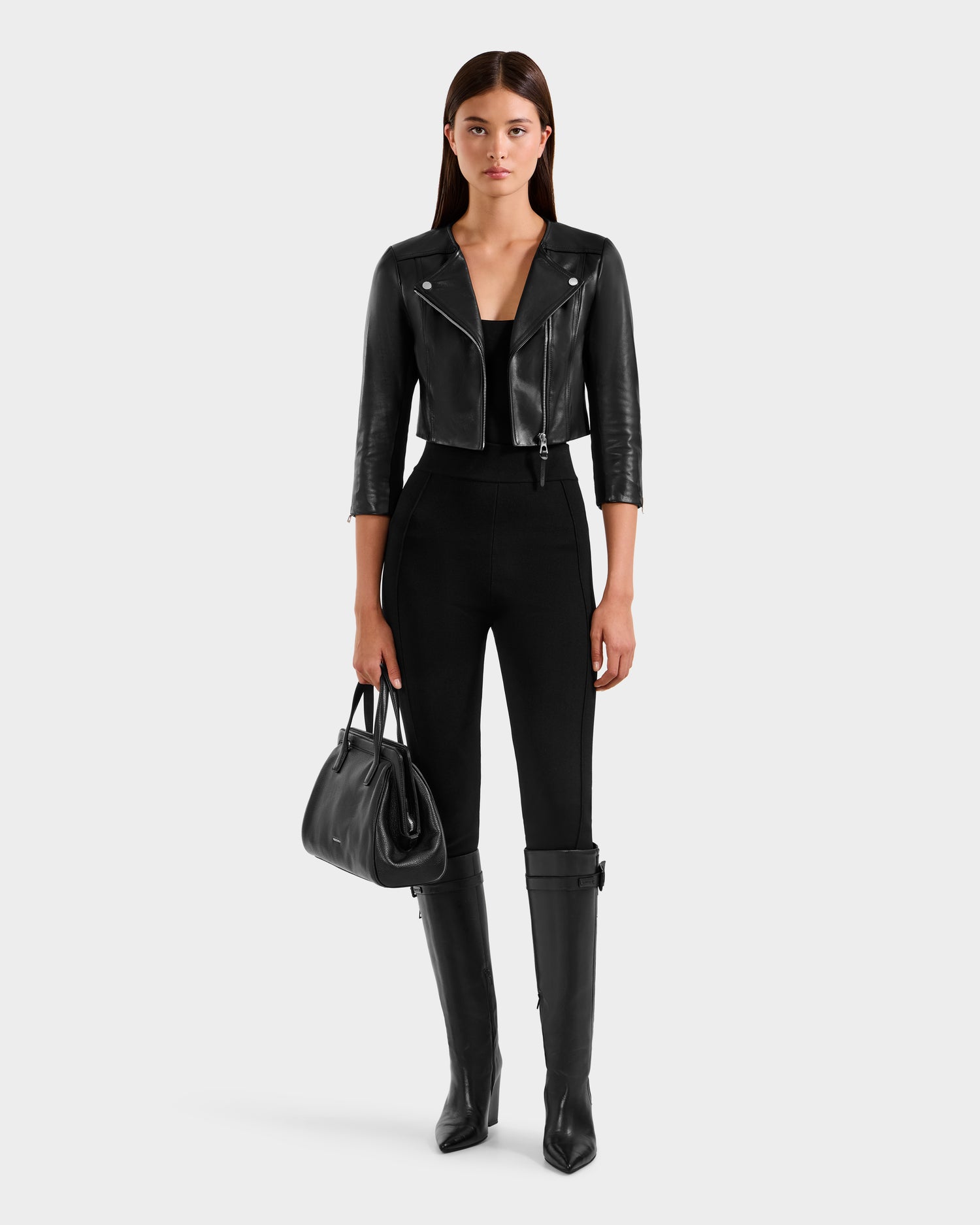A woman with straight brown hair stands facing forward against a white background, wearing the Rudsak WINNI - 8124053 Black cropped lamb leather biker jacket, a fitted black top, high-waisted black pants, knee-high boots, and holding a black handbag.