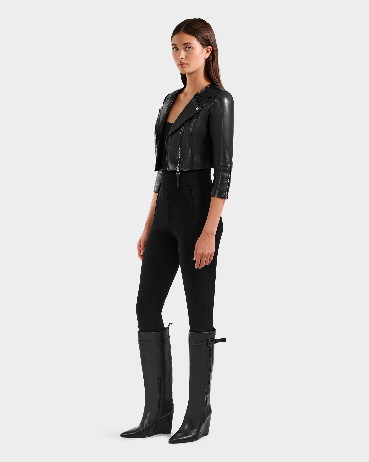 A woman poses against a plain background in the Rudsak WINNI - 8124053 Black cropped biker jacket crafted from soft lamb leather, styled with a black top, fitted pants, and knee-high boots for a chic modern look.