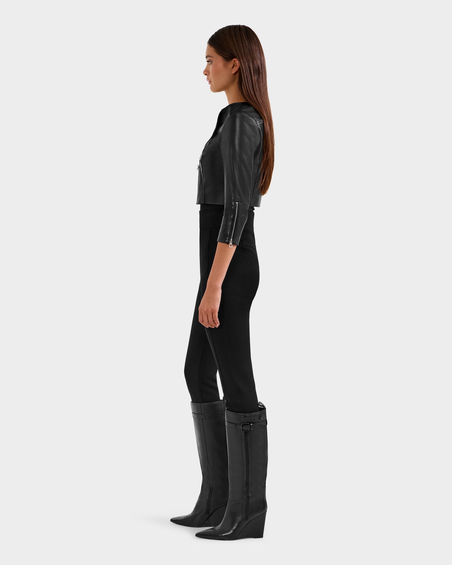 A woman stands in profile against a light gray background, wearing the Rudsak WINNI - 8124053 Black cropped leather biker jacket with black fitted pants and knee-high heeled boots—a chic statement in women's fashion.