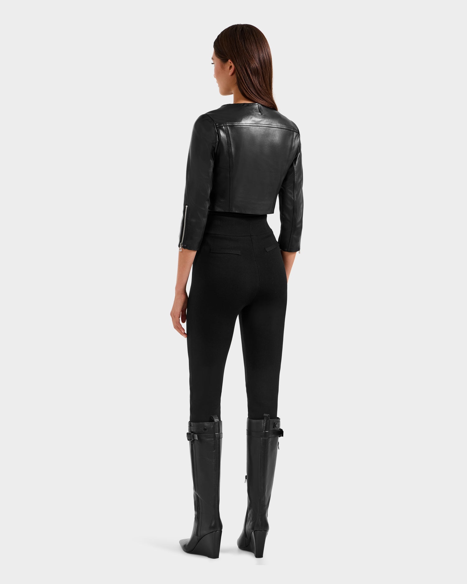 A woman faces away, wearing the Rudsak WINNI - 8124053 Black cropped lamb leather biker jacket, styled with high-waisted black pants and knee-high boots against a plain light gray background—a chic women's fashion look.
