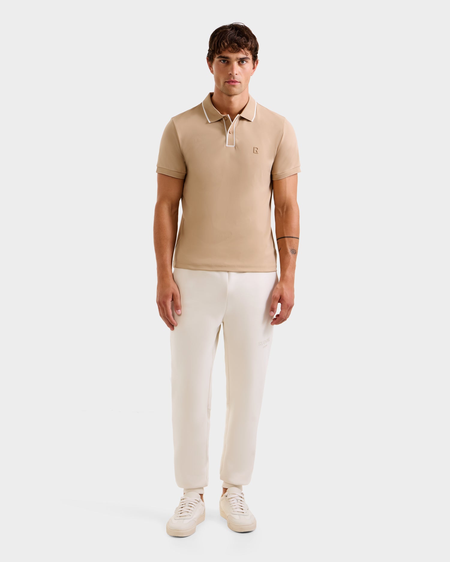 A man stands facing forward, wearing the Rudsak XAVIER - 6524027 Almond polo shirt, paired with white pants and sneakers, against a plain white background.