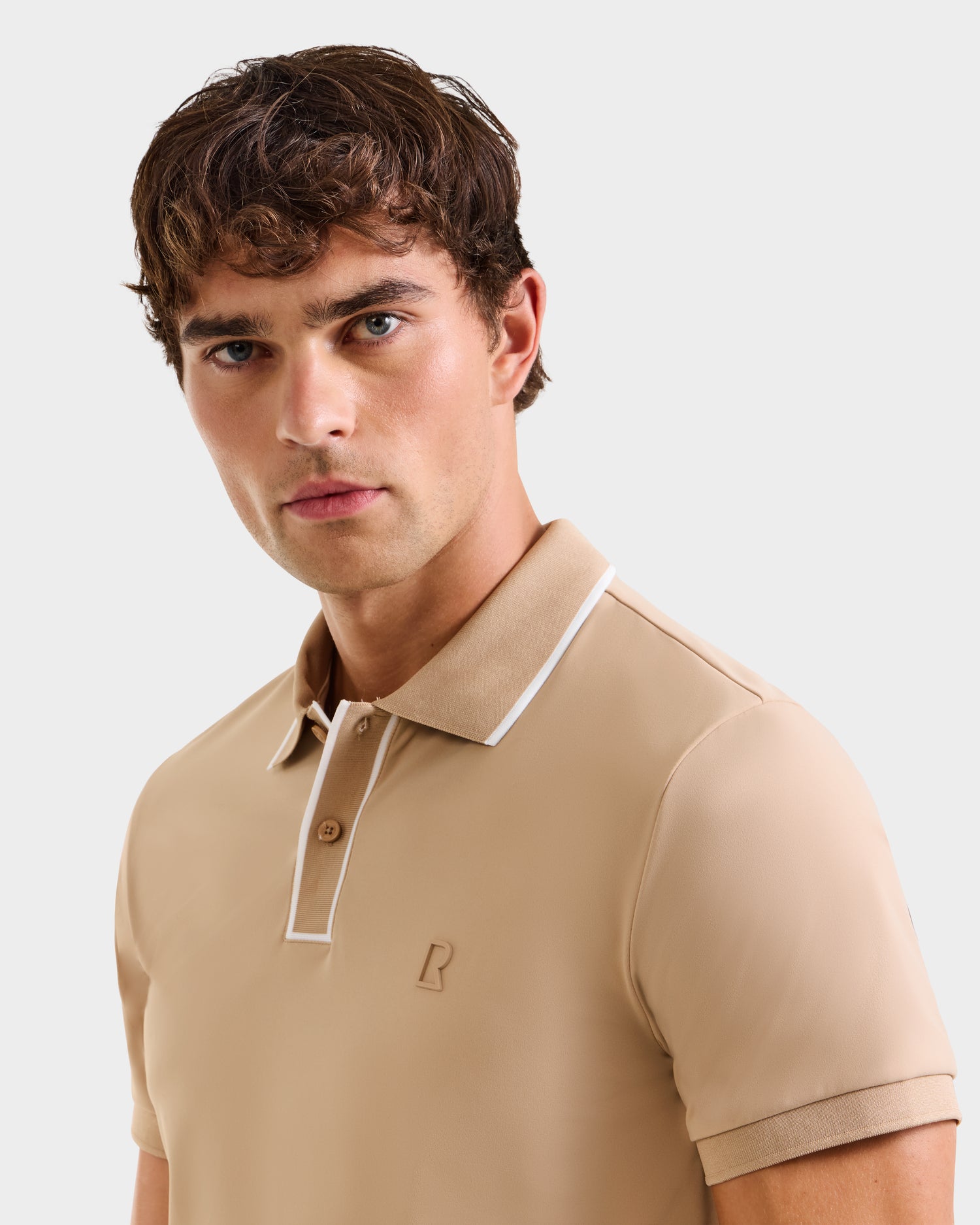 A young man with short brown hair models the Rudsak XAVIER - 6524027 Almond mens polo shirt, featuring white collar trim, standing against a plain white background and looking directly at the camera.