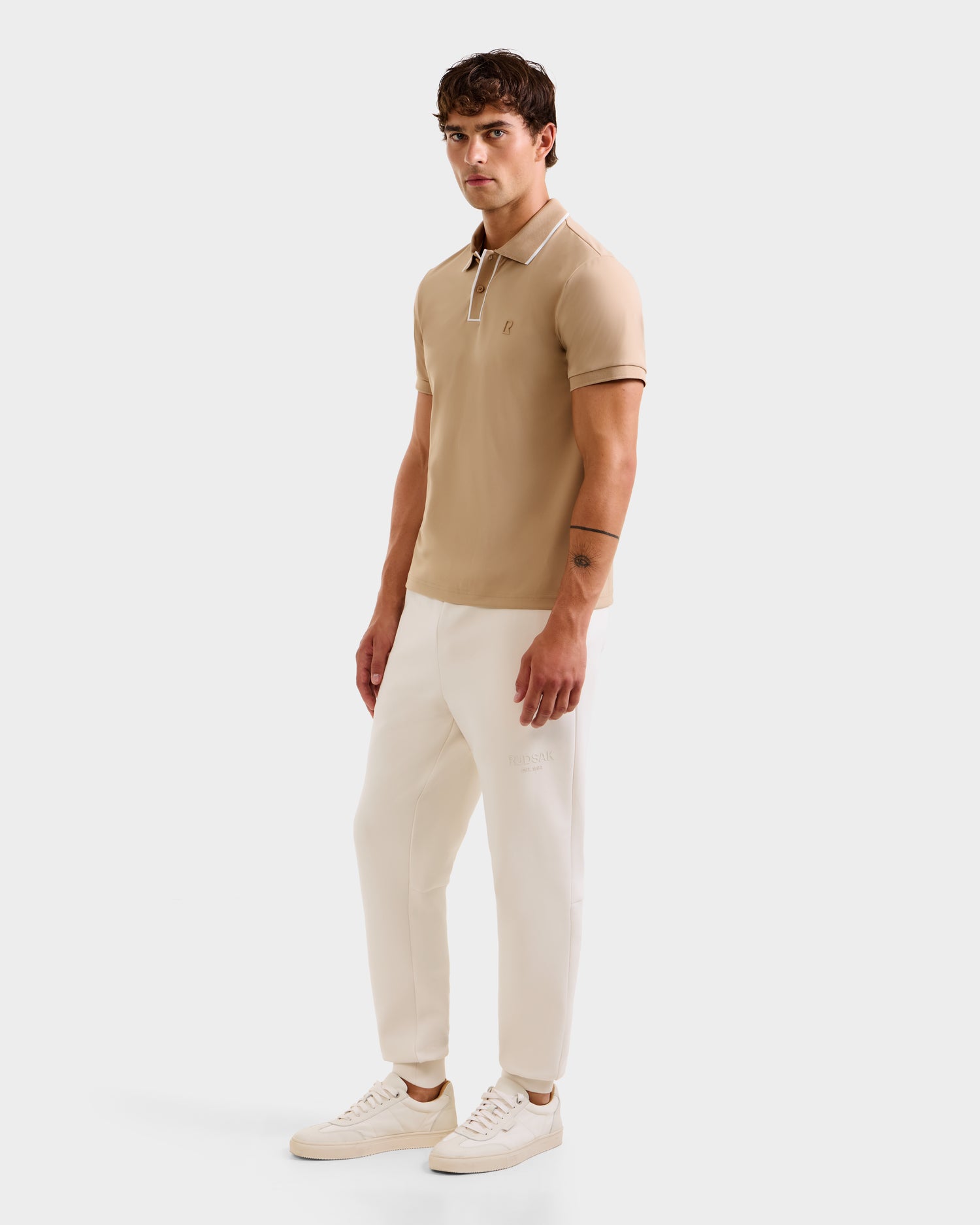 A man with short brown hair and a neutral expression stands against a white background, wearing the Rudsak XAVIER - 6524027 Almond classic polo shirt, cream-colored pants, and white sneakers, one arm relaxed at his side.