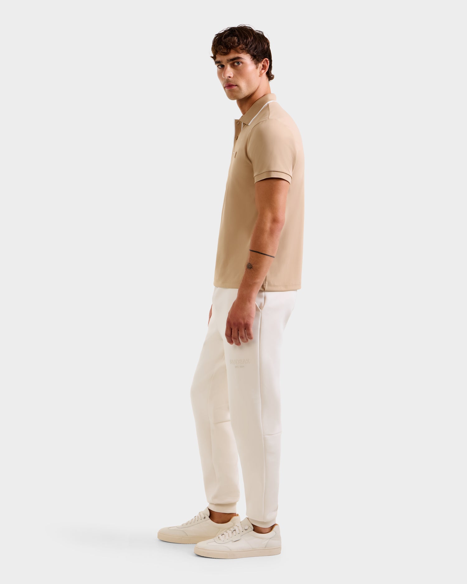 A man with short dark hair and a left forearm tattoo models the Rudsak XAVIER - 6524027 Almond polo shirt, paired with white pants and sneakers, standing sideways against a plain white background.
