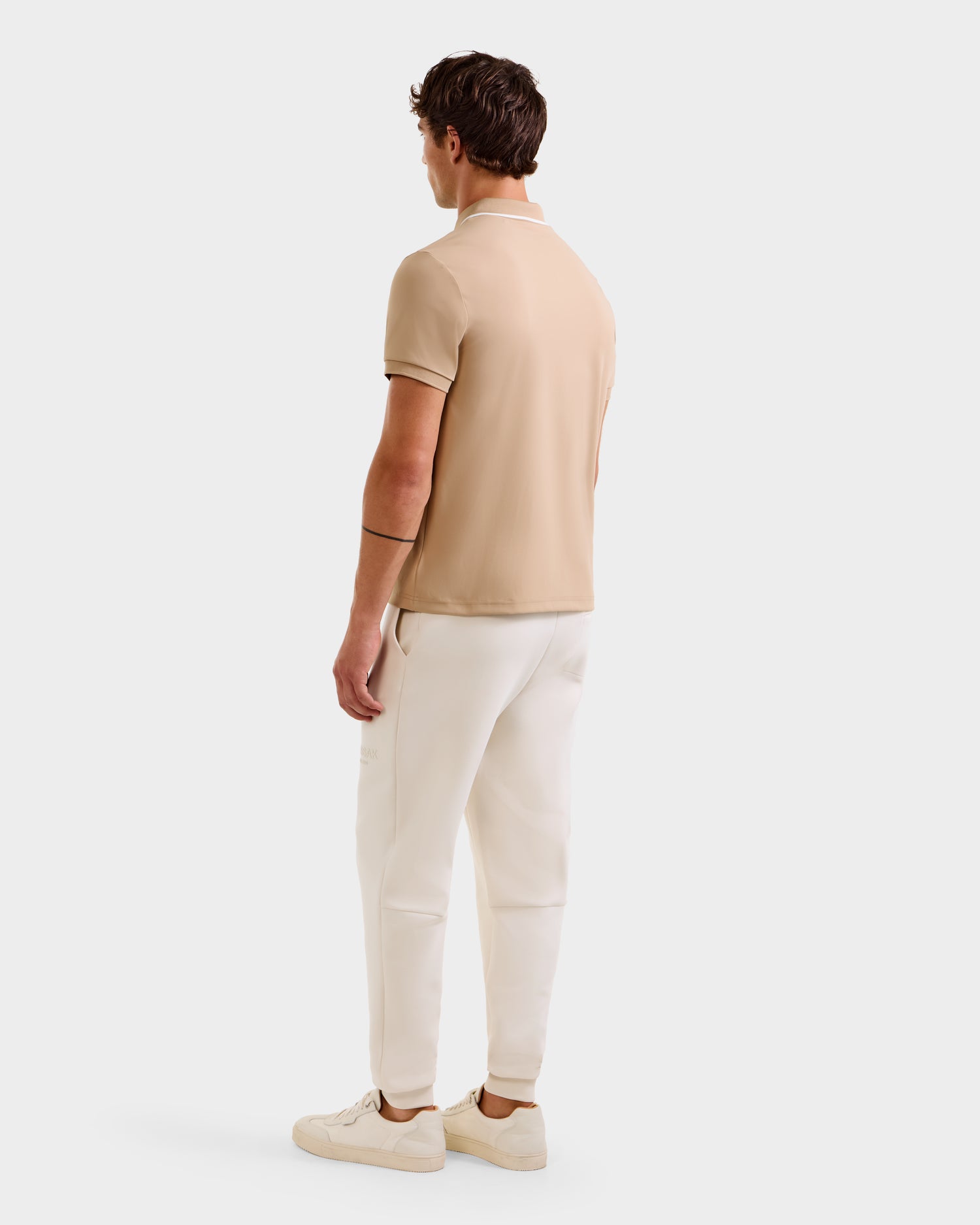 A man faces away, wearing a Rudsak XAVIER - 6524027 Almond polo, off-white pants, and beige sneakers against a plain white background.