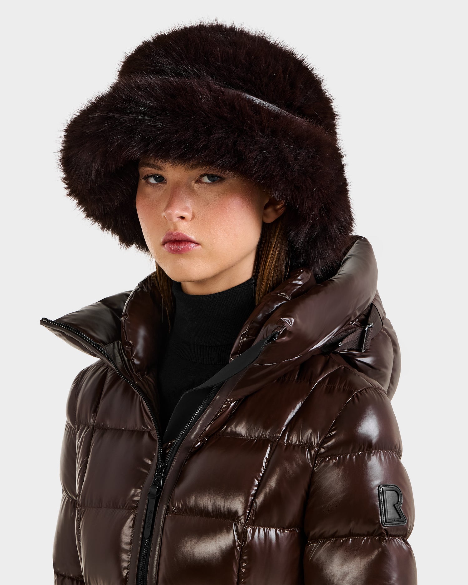 Wearing the Rudsak YURI - L425538 Espresso puffer jacket with vegan-leather trim and a faux-fur bucket hat, a woman gazes ahead with a neutral expression, showcasing chic women’s winter style against a plain background.