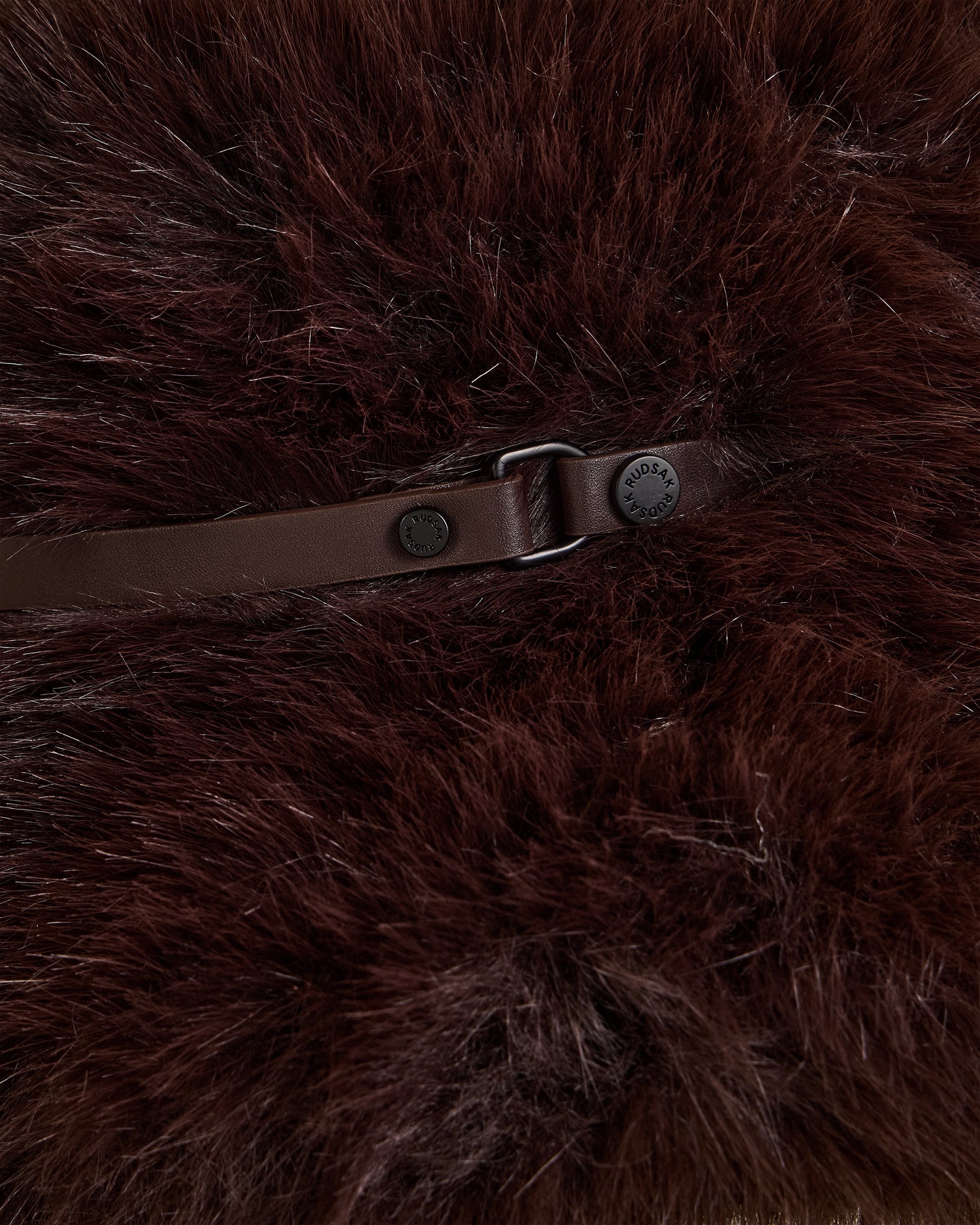 A close-up of Rudsak's YURI - L425538 Espresso shows dark brown faux fur with lighter streaks, accented by a vegan-leather strap and inscribed metal buckle—details of this women's winter accessory.