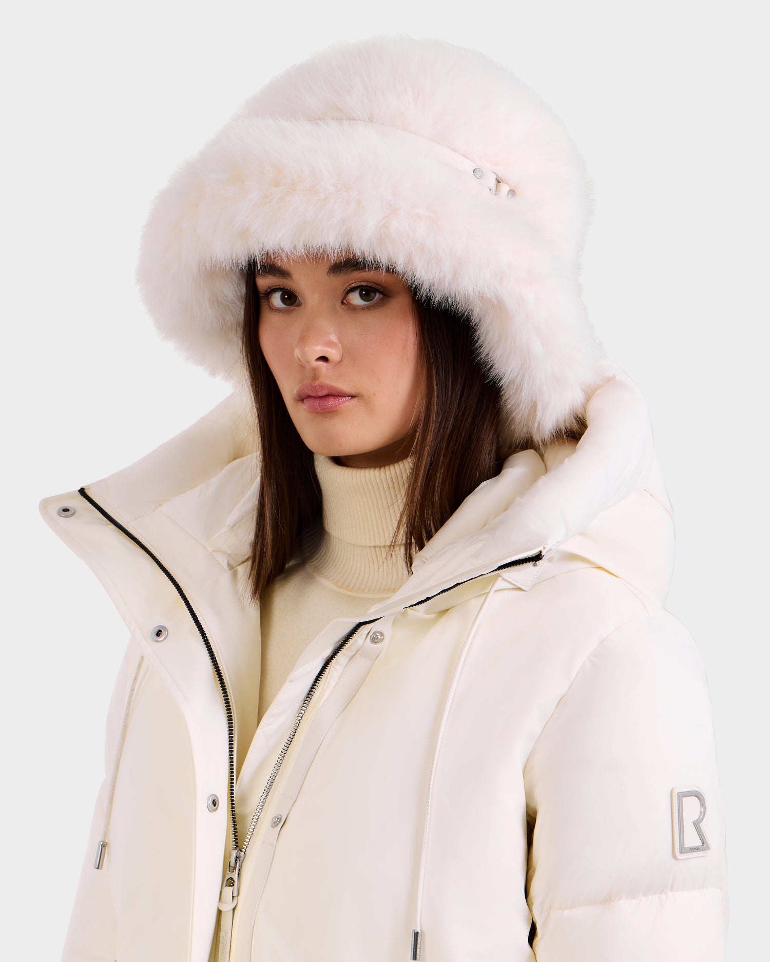 A woman wears the Rudsak YURI - L425538 Iceland white puffer jacket with a high collar and a fluffy faux-fur bucket hat, her straight brown hair and neutral expression complementing this chic winter accessory against a light background.