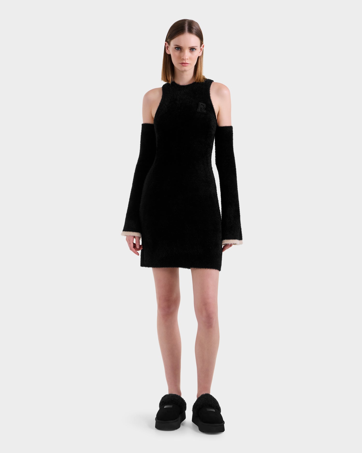 A woman faces forward in the Rudsak ZARAH - 8525696 Black dress, featuring cold shoulder cutouts and matching detached sleeves. With straight, shoulder-length hair and black platform shoes, she channels après-ski style against a plain light gray background.