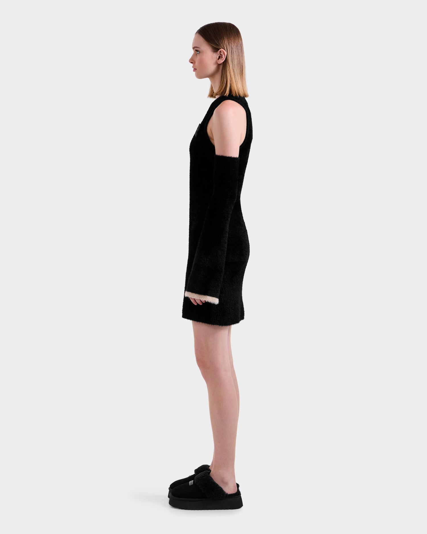 A woman stands in profile against a plain background, wearing the Rudsak ZARAH - 8525696 Black fitted dress with matching long black gloves and black platform shoes. Her straight hair is center-parted and falls to her shoulders.