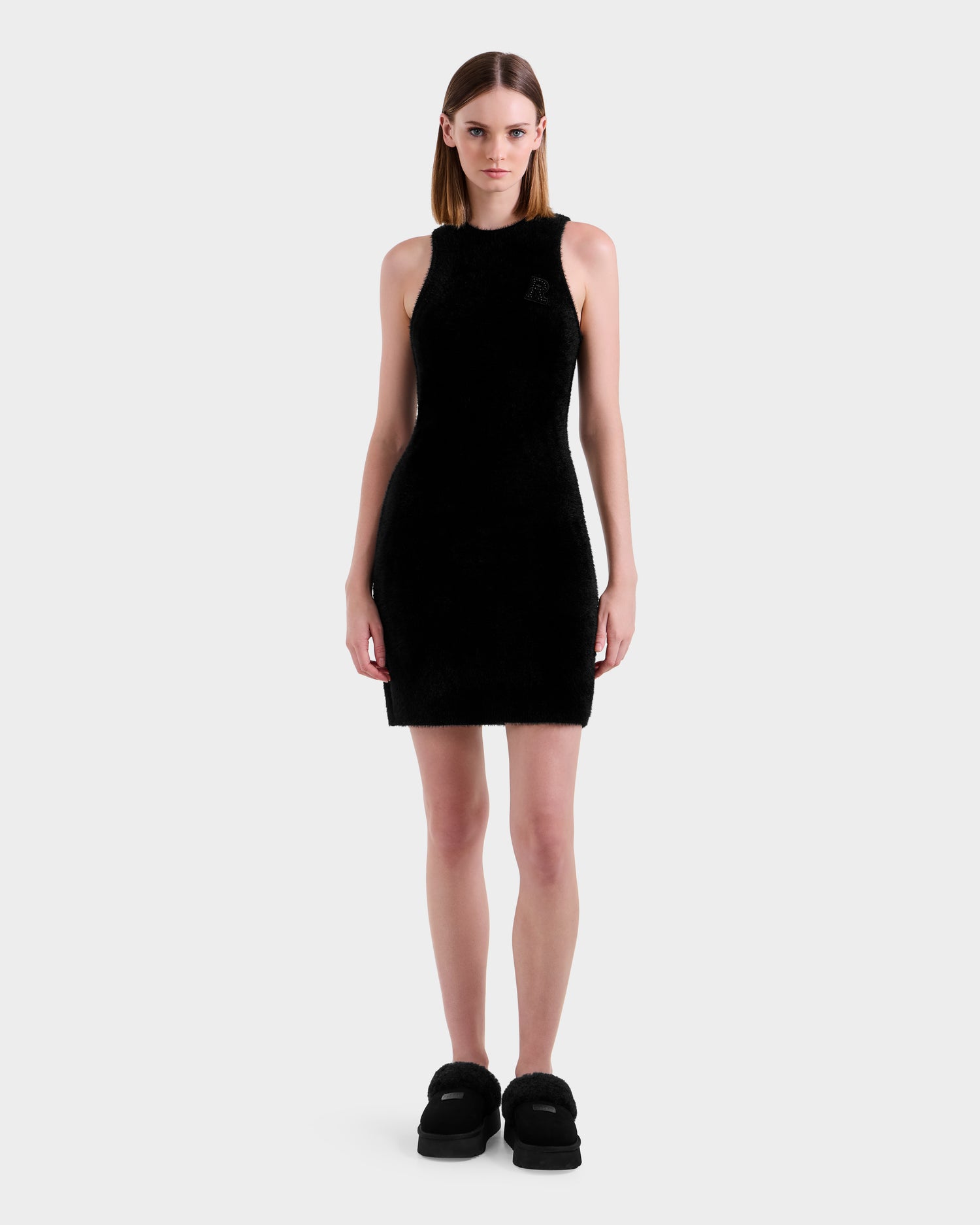 A woman with straight brown hair wears the Rudsak ZARAH - 8525696 Black dress, paired with black platform shoes, standing against a plain light gray background and facing forward with a neutral expression.
