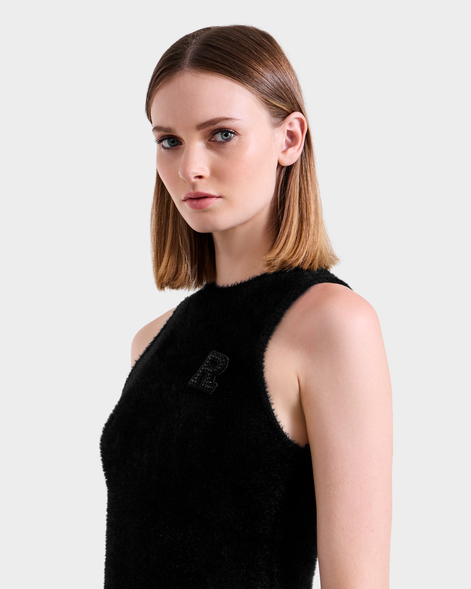 A woman with straight, shoulder-length light brown hair models the ZARAH - 8525696 Black sleeveless fuzzy knit dress by Rudsak, featuring a small R emblem on the left side, against a plain light gray background.