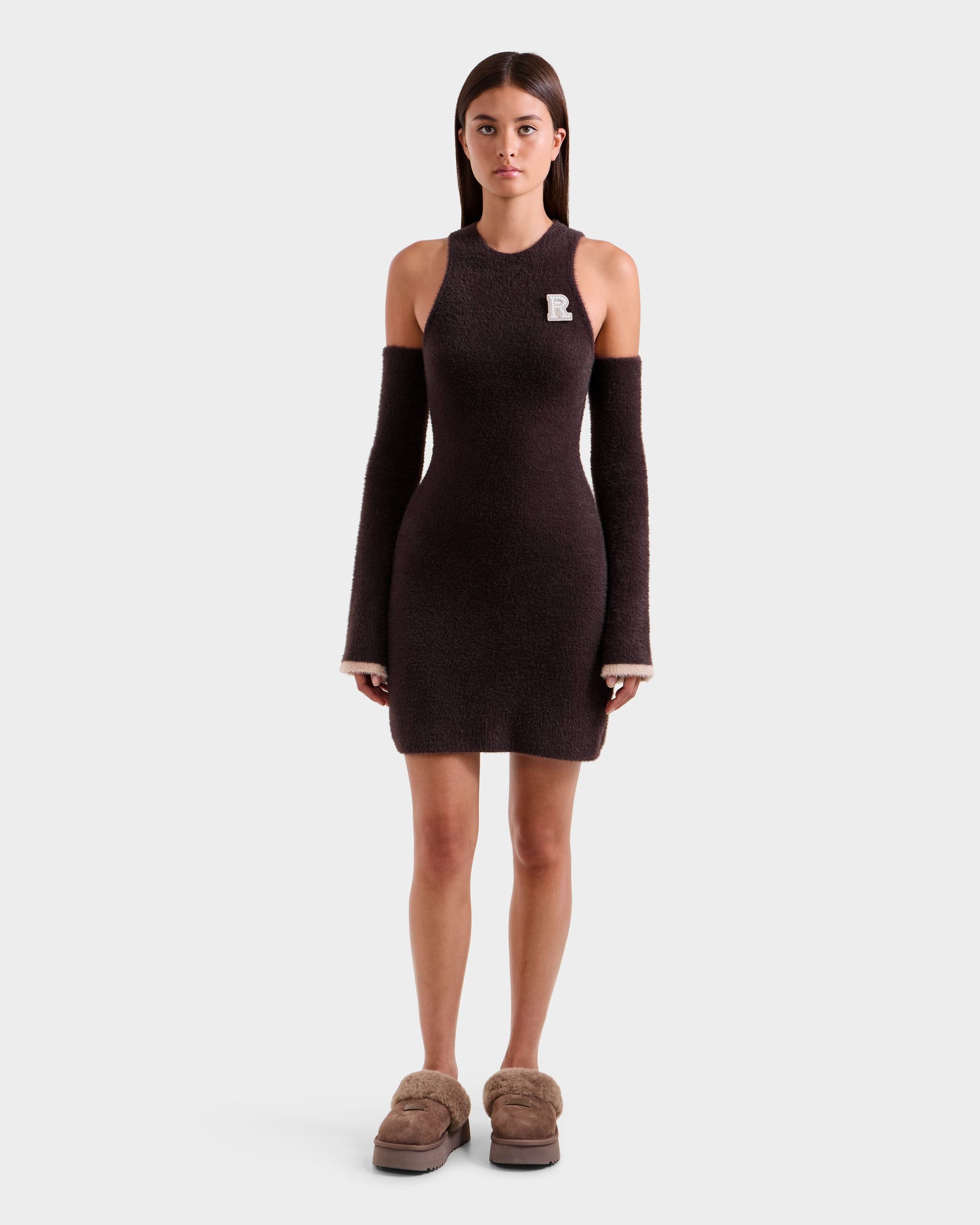 A woman faces forward in the ZARAH - 8525696 Espresso dress by Rudsak, featuring matching detached sleeves, plush beige platform slippers, and a small R patch on the upper left, highlighting its versatile style against a plain background.