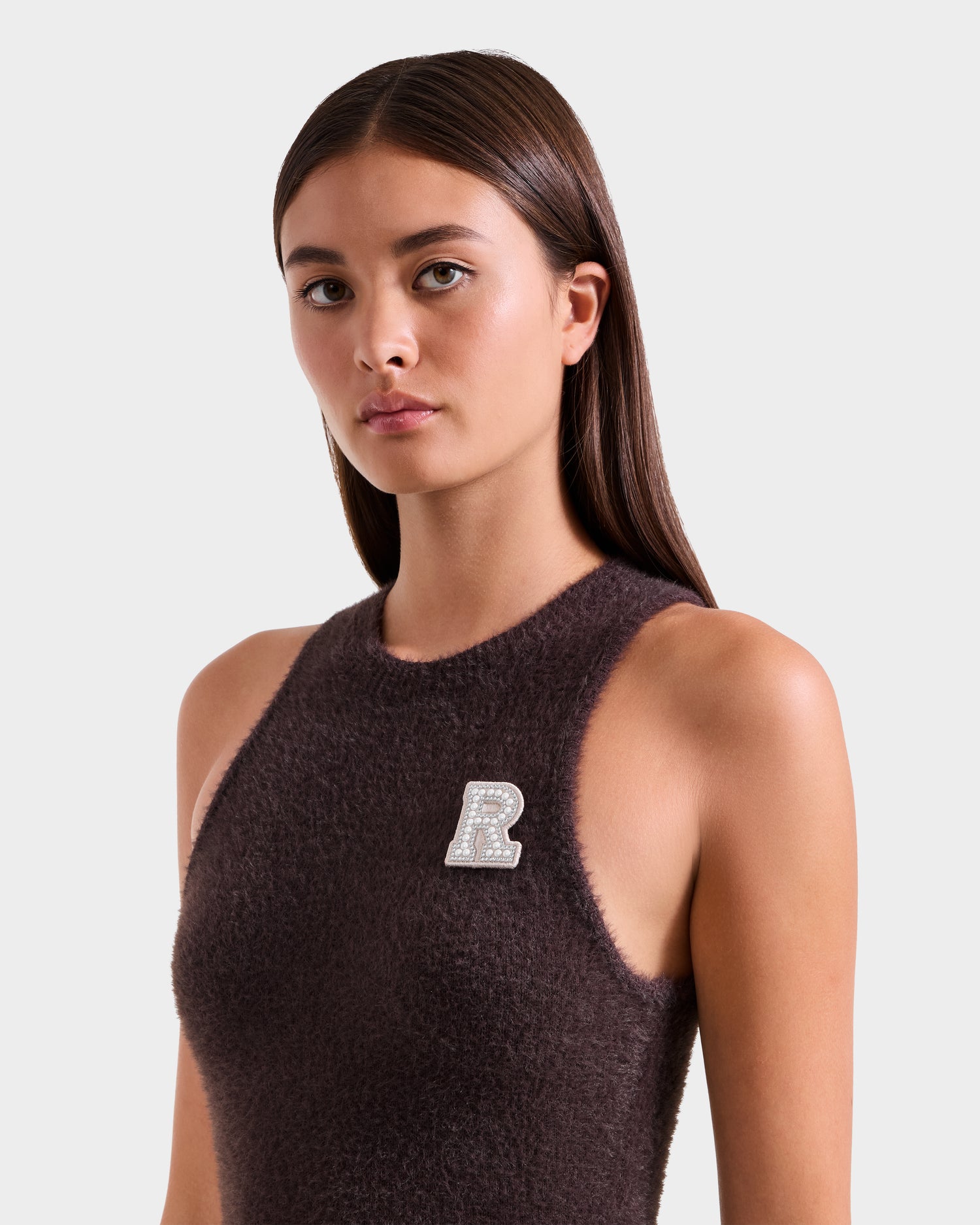 A woman with straight brown hair wears the Rudsak ZARAH - 8525696 Espresso sleeveless textured top, adorned with a rhinestone R brooch on the left, as she poses confidently against a plain light background, looking at the camera.