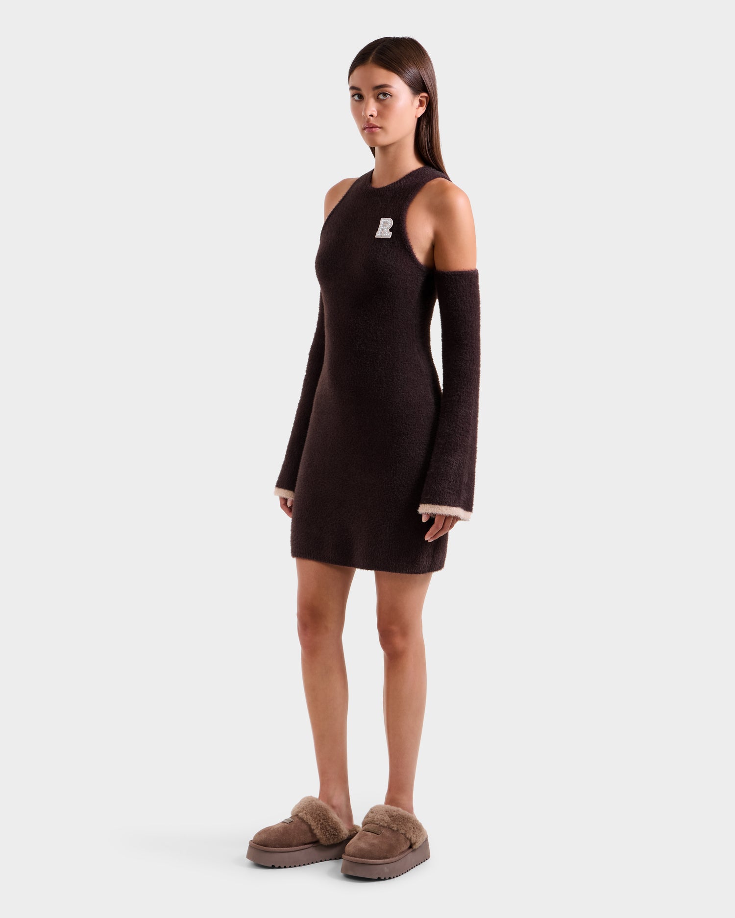 A woman with long straight hair and a neutral expression faces forward, wearing the Rudsak ZARAH - 8525696 Espresso one-shoulder women's fitted dress with detached long sleeves and tan fuzzy slippers, against a plain white background.