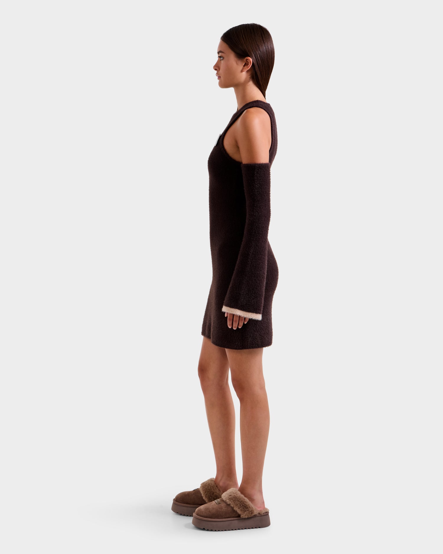 A woman stands in profile wearing the Rudsak ZARAH - 8525696 Espresso women's fitted knit dress with matching detached sleeves and tan fuzzy slippers, showcasing versatile styling against a plain light gray background.
