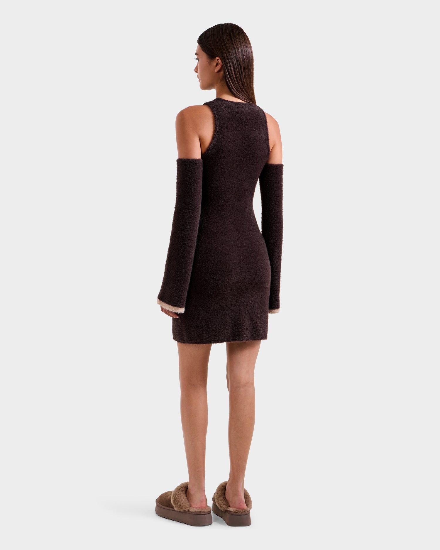 A woman stands facing away, wearing the Rudsak ZARAH - 8525696 Espresso sleeveless fitted dress with matching detached long sleeves and brown fuzzy slippers, set against a plain light background.