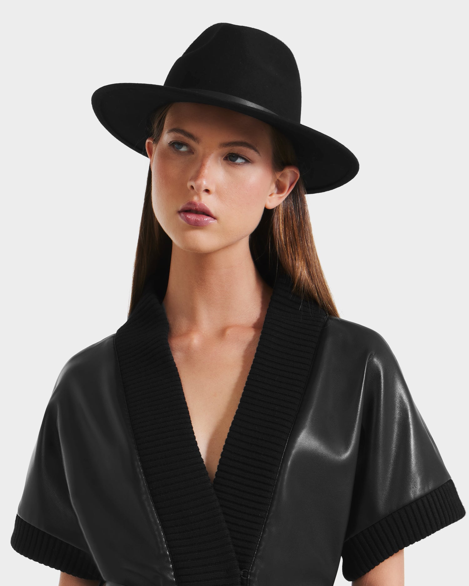 A woman with straight brown hair wears the Rudsak ZELDA - 8425536 Black short-sleeved, leather-textured jacket with a ribbed collar and a wide-brim hat, posing against a plain light background with a neutral expression.