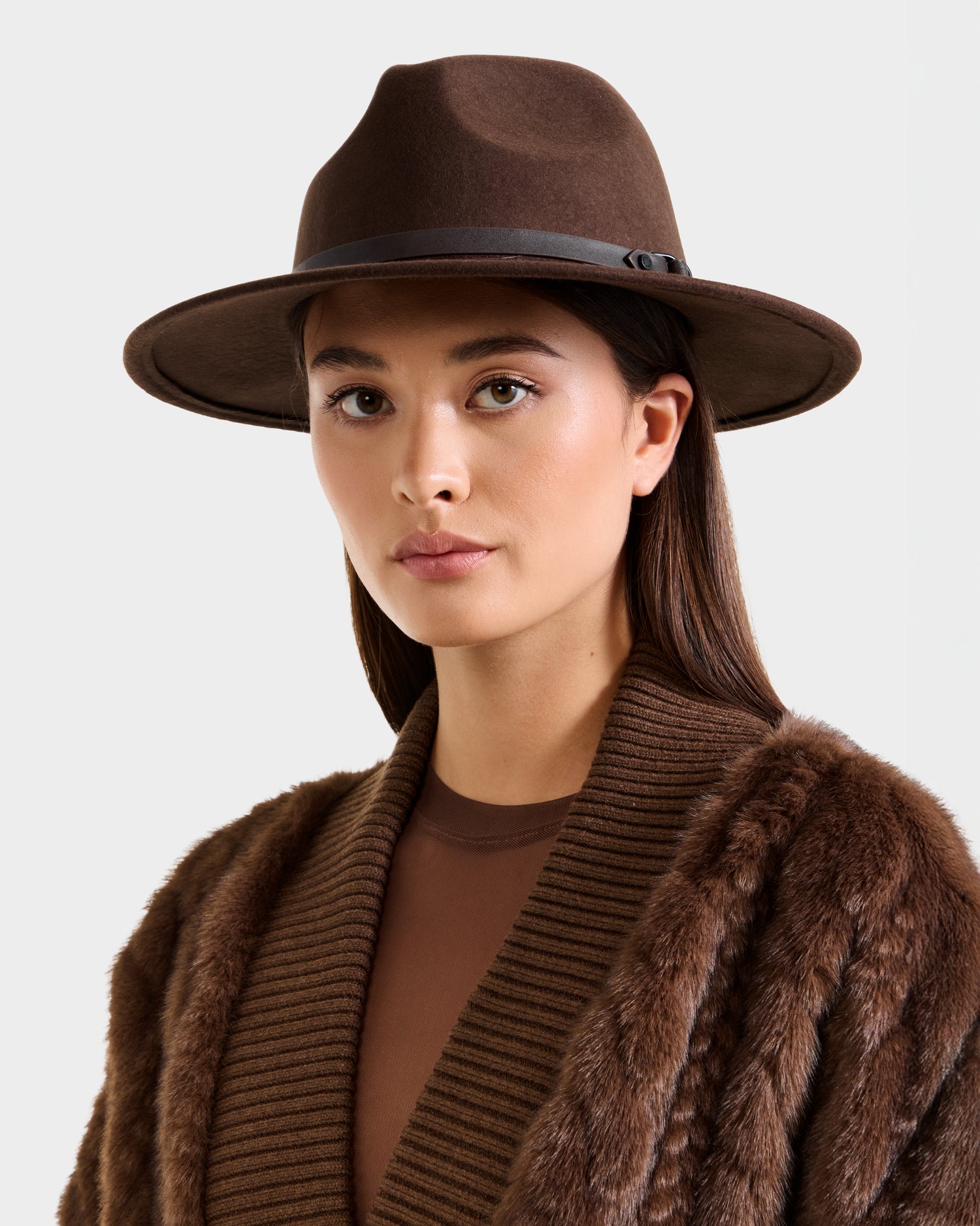 A woman models the Rudsak ZELDA - 8425536 Espresso fur coat, paired with a wide-brim hat, gazing directly at the camera against a plain light background.