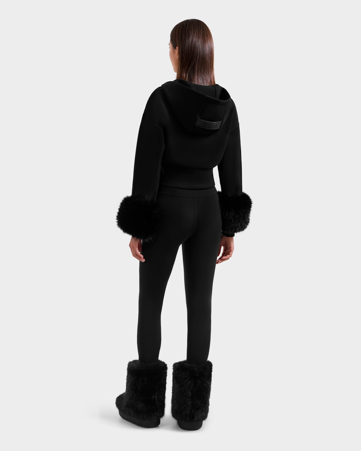 A woman with straight brown hair faces away, wearing the Rudsak ZILLIA - 8525673 Black women’s cropped hoodie with faux fur cuffs, along with matching black leggings and black fur boots against a plain light gray background.