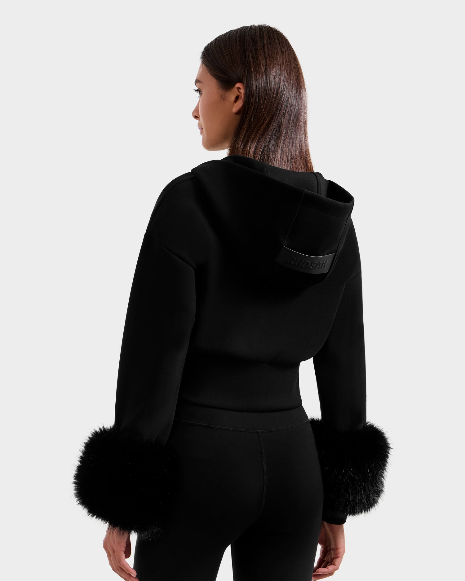 A woman with straight brown hair, seen from the back, wears Rudsak's ZILLIA - 8525673 Black cropped hoodie with faux fur cuffs and matching black pants, standing against a plain light background.