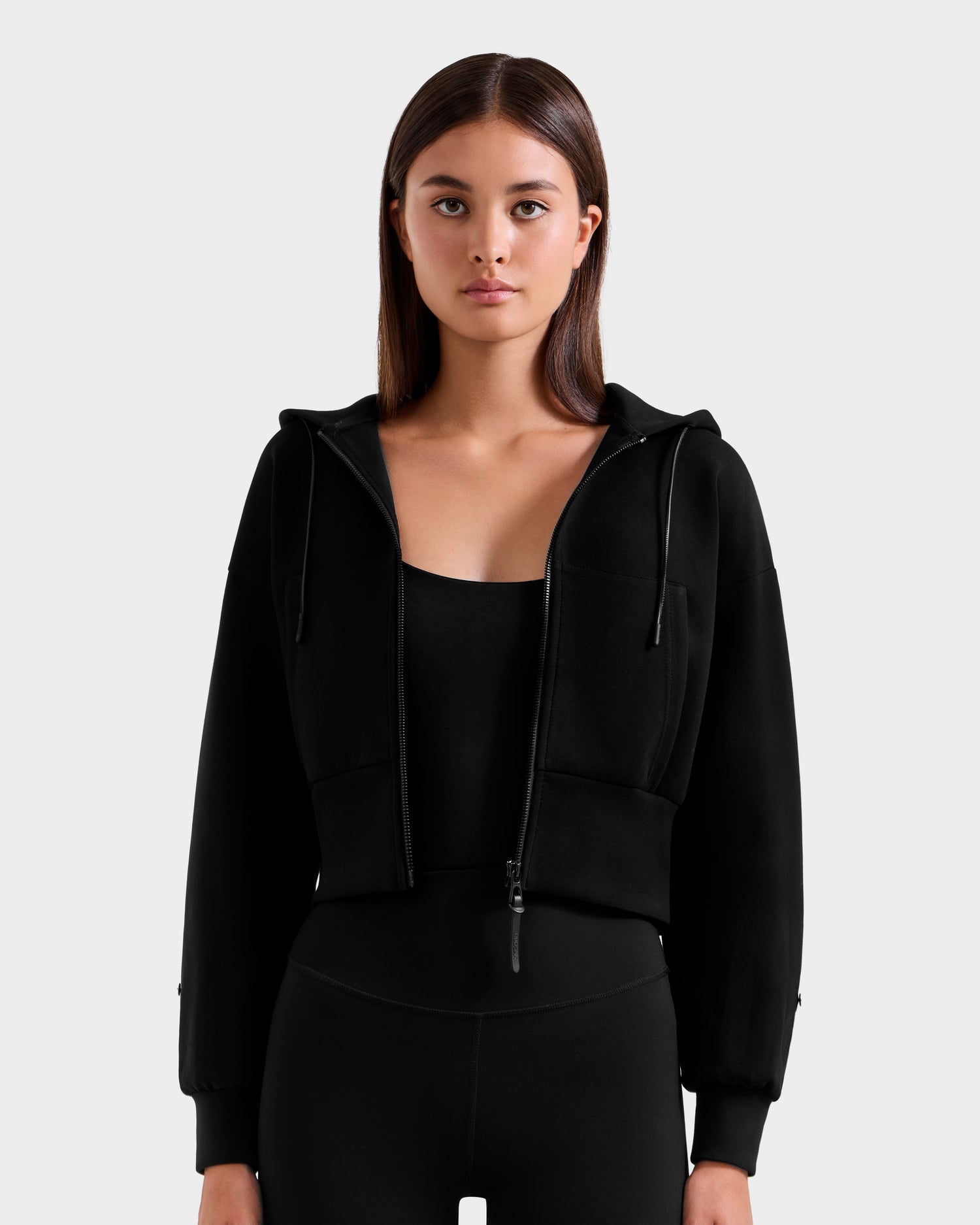 A woman with straight brown hair wears the Rudsak ZILLIA - 8525673 Black women’s cropped hoodie with faux fur cuffs over a black top and leggings, standing against a plain light gray background, facing forward with a neutral expression.