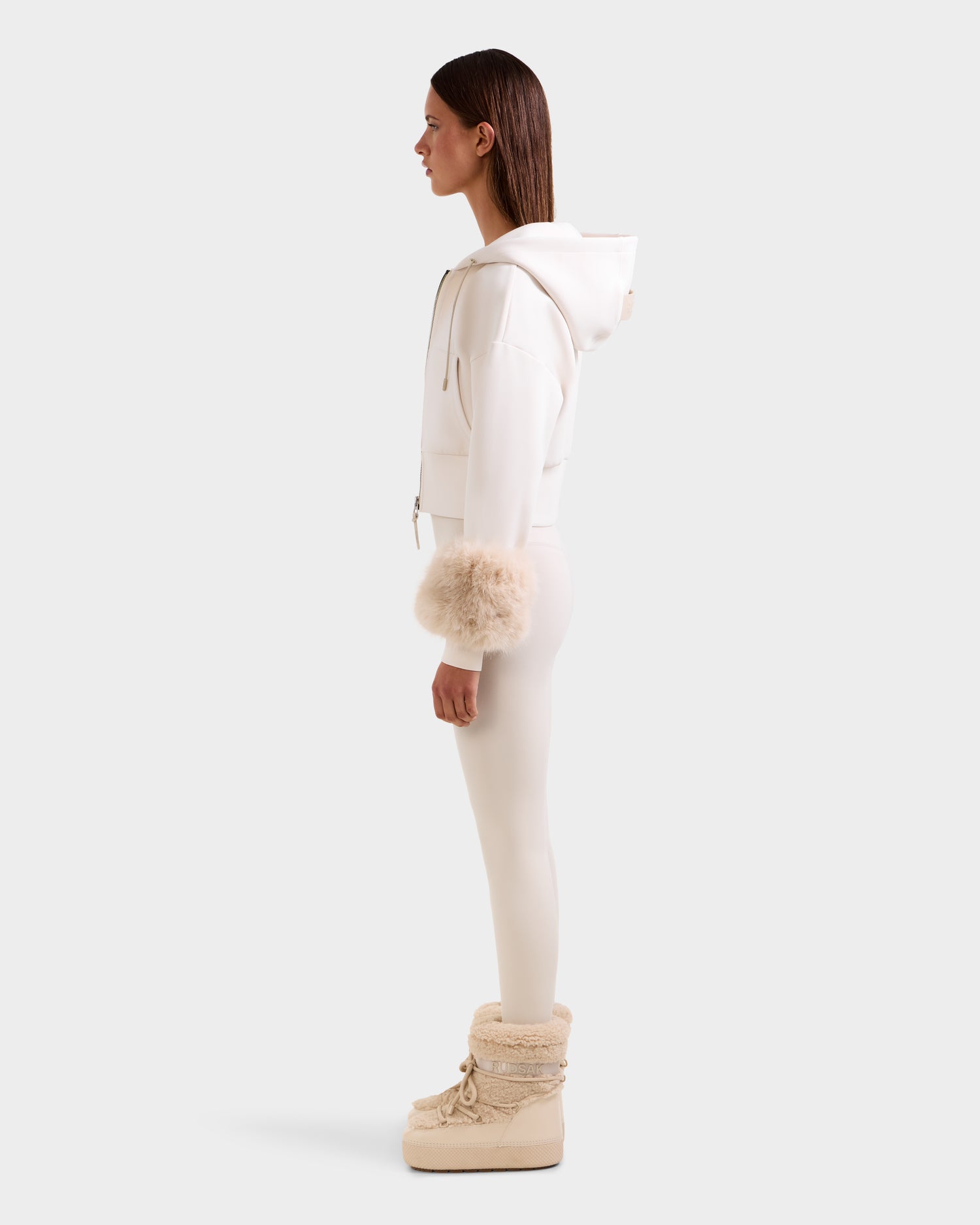 A person stands in profile against a plain light background, wearing the Rudsak ZILLIA - 8525673 Vanilla women’s hoodie with faux fur wrist cuffs, fitted leggings, and beige fur-trimmed winter boots.