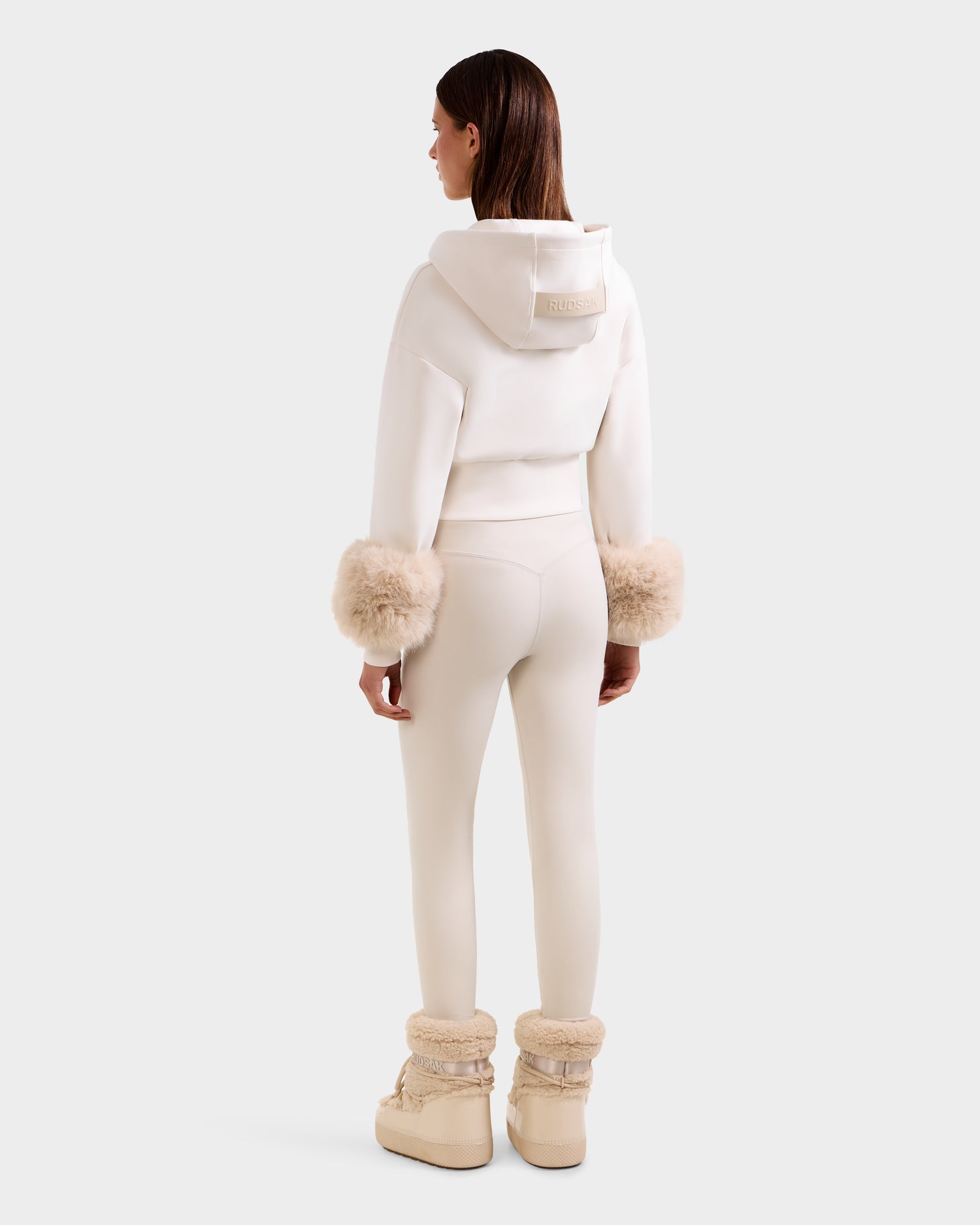 A woman with straight brown hair faces away, wearing the Rudsak ZILLIA - 8525673 Vanilla off-white hoodie with faux fur wrist cuffs, matching leggings, and beige fur-trimmed boots against a plain white background.