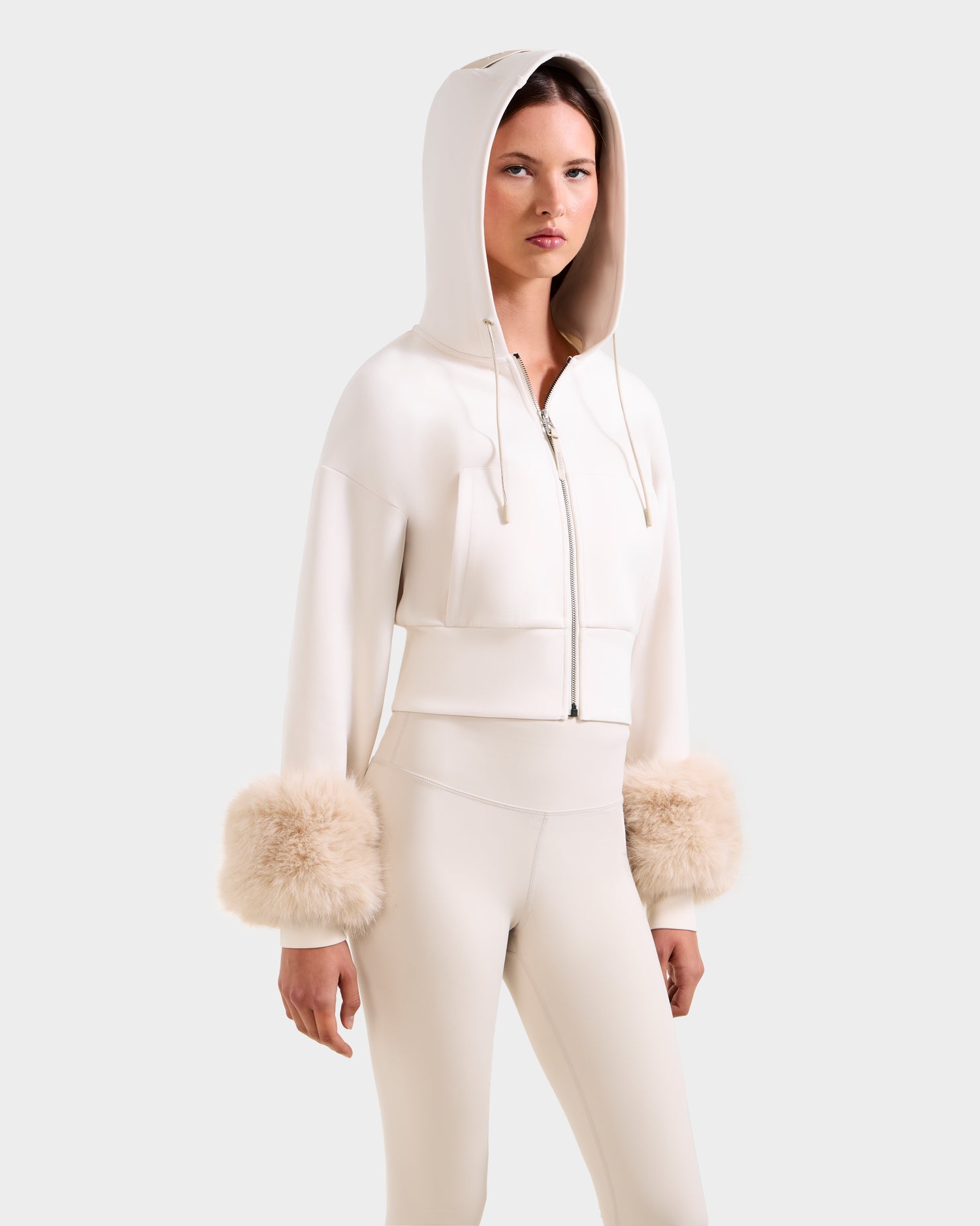 A woman models the Rudsak ZILLIA - 8525673 Vanilla cream-colored cropped zip-up hoodie with faux fur cuffs and matching high-waisted pants against a light gray background.