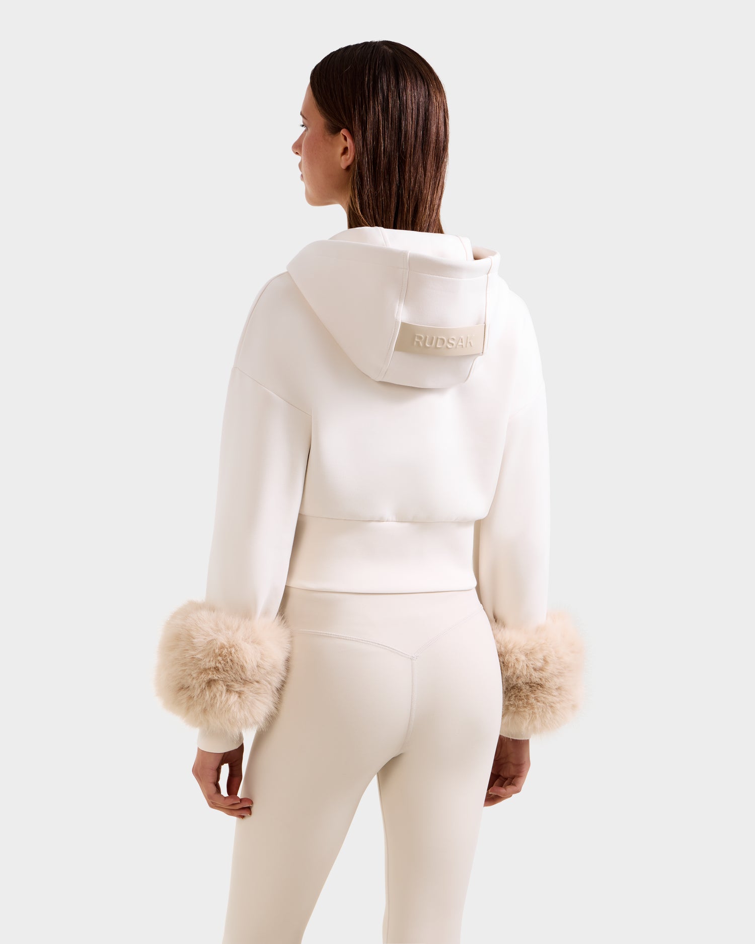 A woman wears the Rudsak ZILLIA – 8525673 Vanilla cream cropped zip-up hoodie with faux fur wrist cuffs and fitted pants, viewed from the back; "RUDSAK" is printed on the hoodie’s hood.