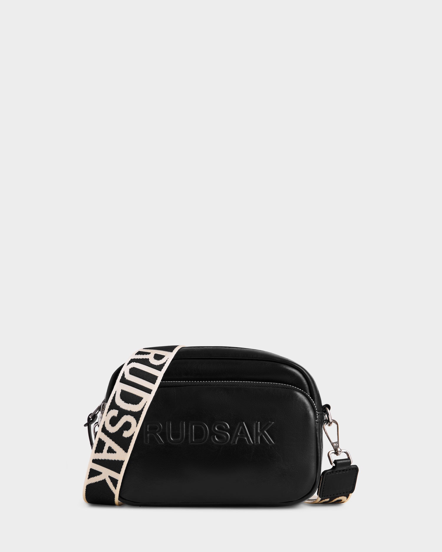 The ZOYA L - 8325543 Black by Rudsak is a smooth black leather crossbody with RUDSAK embossed on the front, silver hardware, a zippered compartment, and a detachable adjustable beige logo strap with bold black lettering.