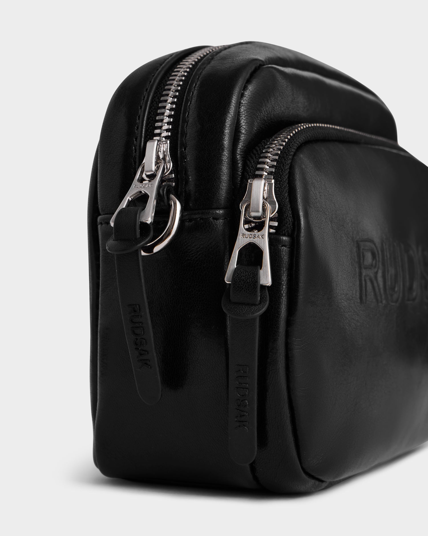 Close-up of the Rudsak ZOYA L - 8325543 Black crossbody bag in smooth black leather, featuring two zippered compartments with silver hardware and embossed pull tabs, plus a sleek minimalist design and adjustable logo strap.