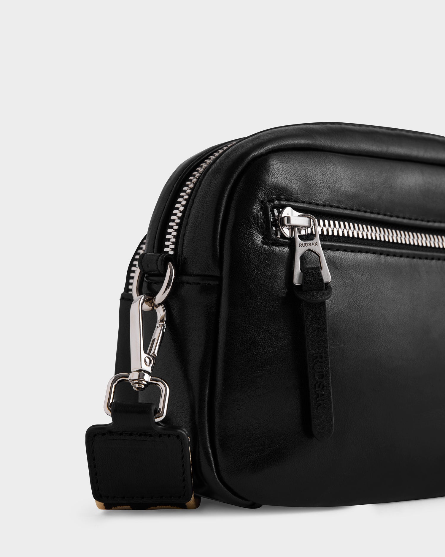 Close-up of the Rudsak ZOYA L - 8325543 black leather crossbody bag, featuring a silver front zipper, branded pull, and adjustable logo strap with metal clasp, set against a plain white background.