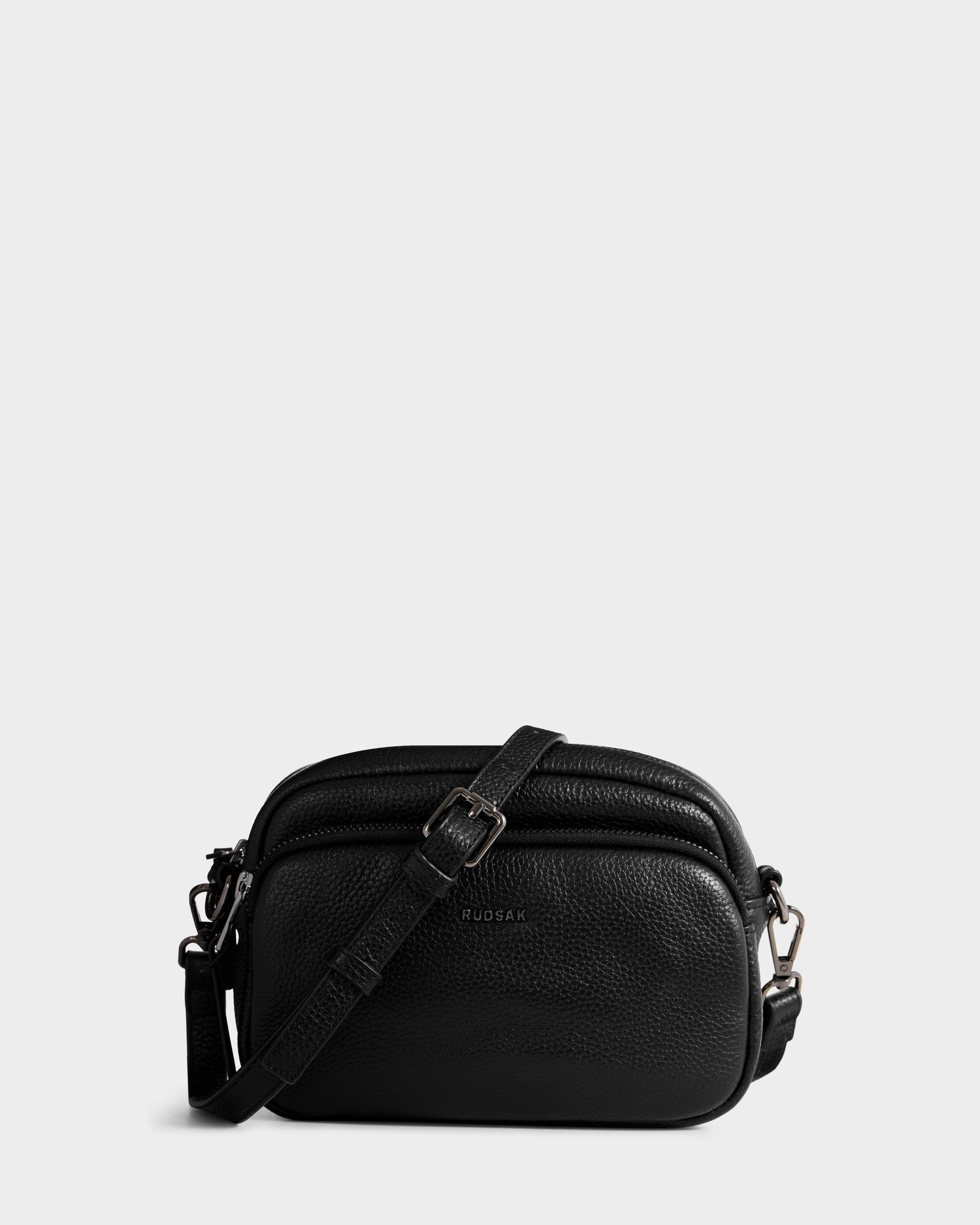 The ZOYA - L325015 Black by Rudsak is a small women’s black leather crossbody bag with an adjustable strap, pebbled texture, and silver hardware. The brand name Rudsak is printed on the front. Displayed on a light gray background.